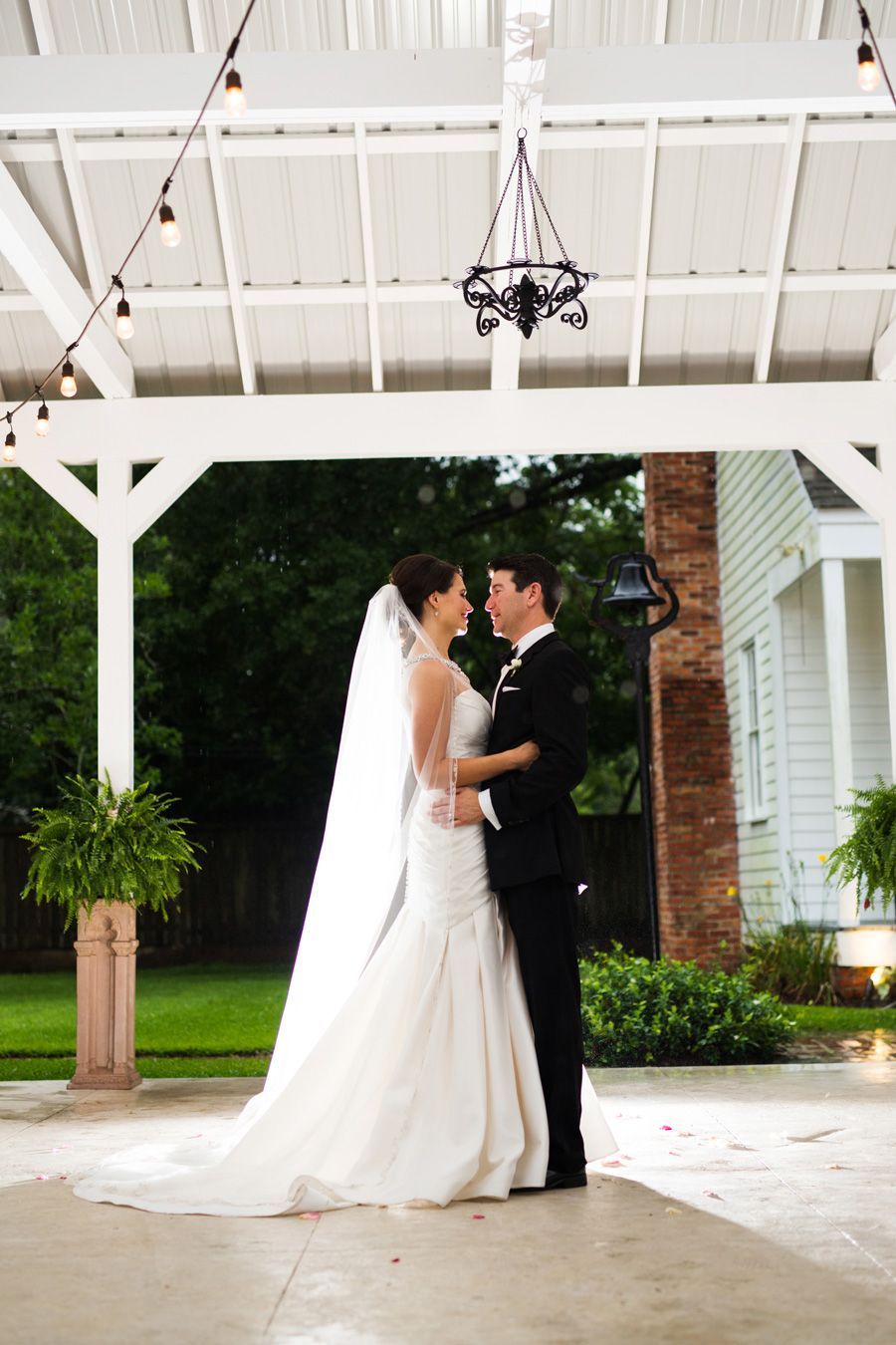 Bride and groom embrace under a white pergola, smiles exchanged, wedding setting.