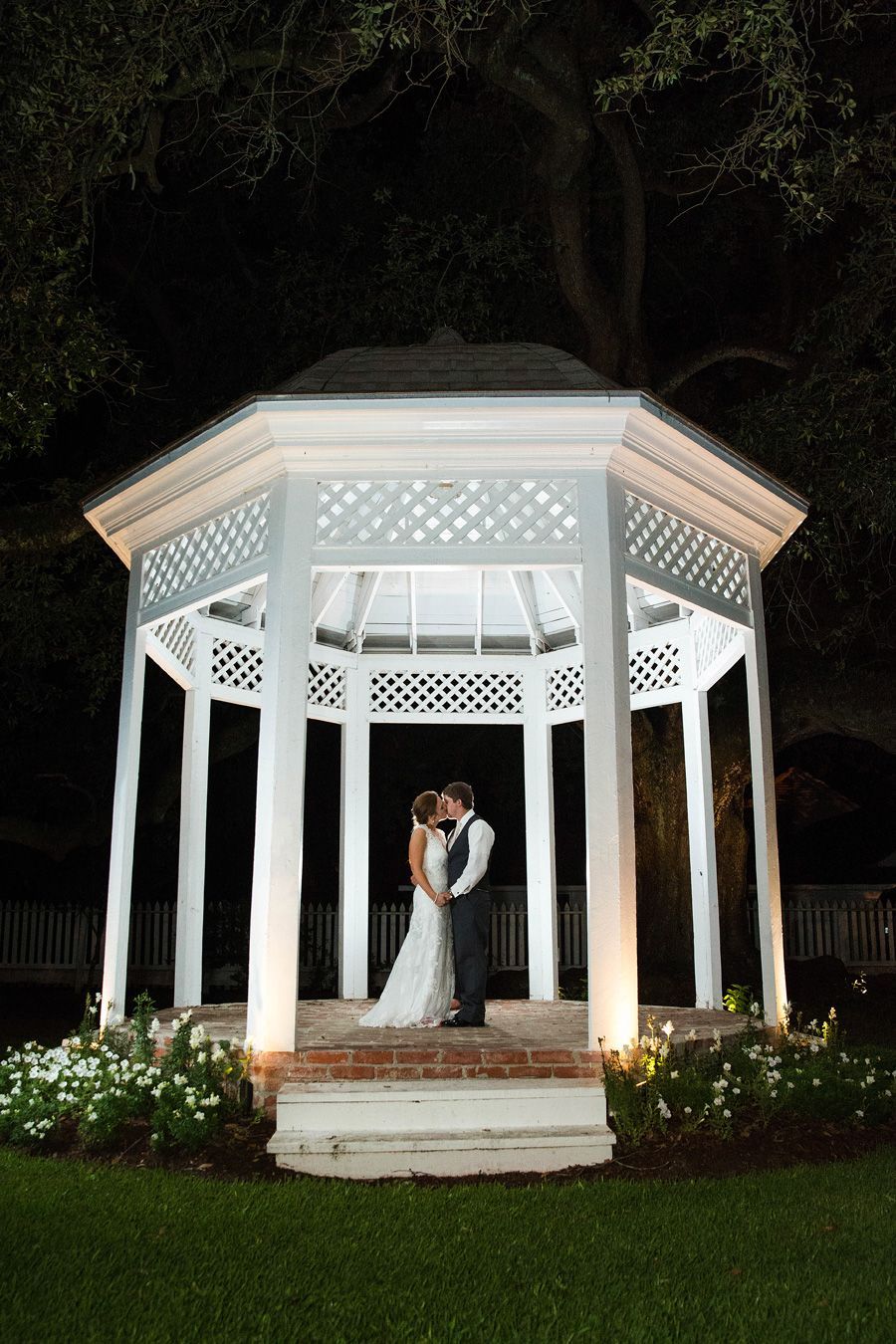 Couple kissing inside a white gazebo at night; lit by lights, surrounded by greenery.
