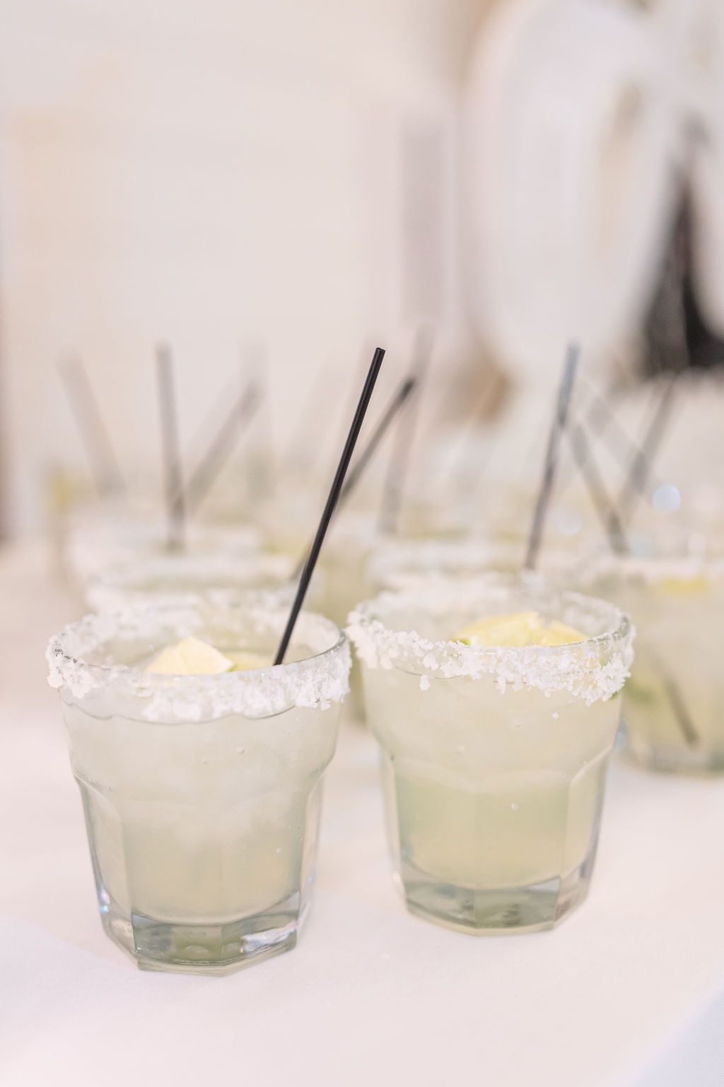 Cocktails in salt-rimmed glasses with black straws. Several are in focus, others blurred. Light background.