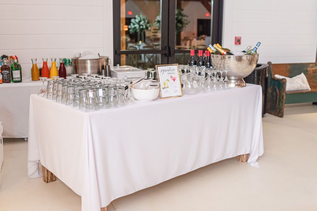 Buffet table with drinks and glasses. White tablecloth, juice bottles, wine, ice bucket, and glasses.