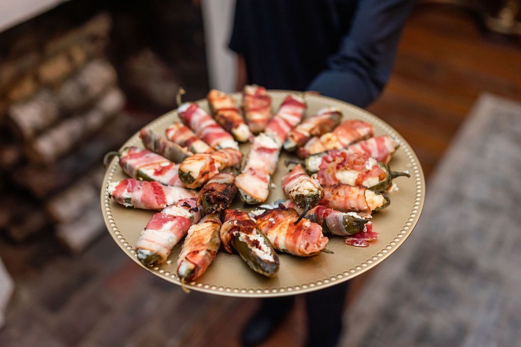 A server holding a gold platter of bacon-wrapped jalapeño poppers near a fireplace.