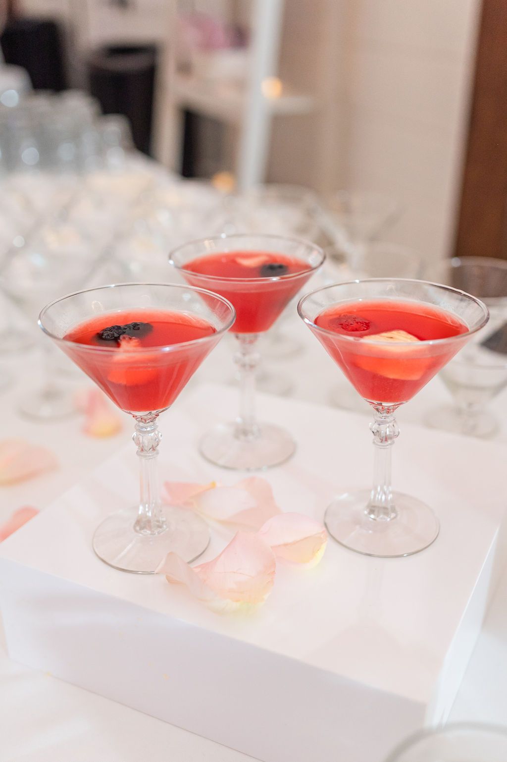 Three red cocktails with fruit in martini glasses, on a white surface, with rose petals.