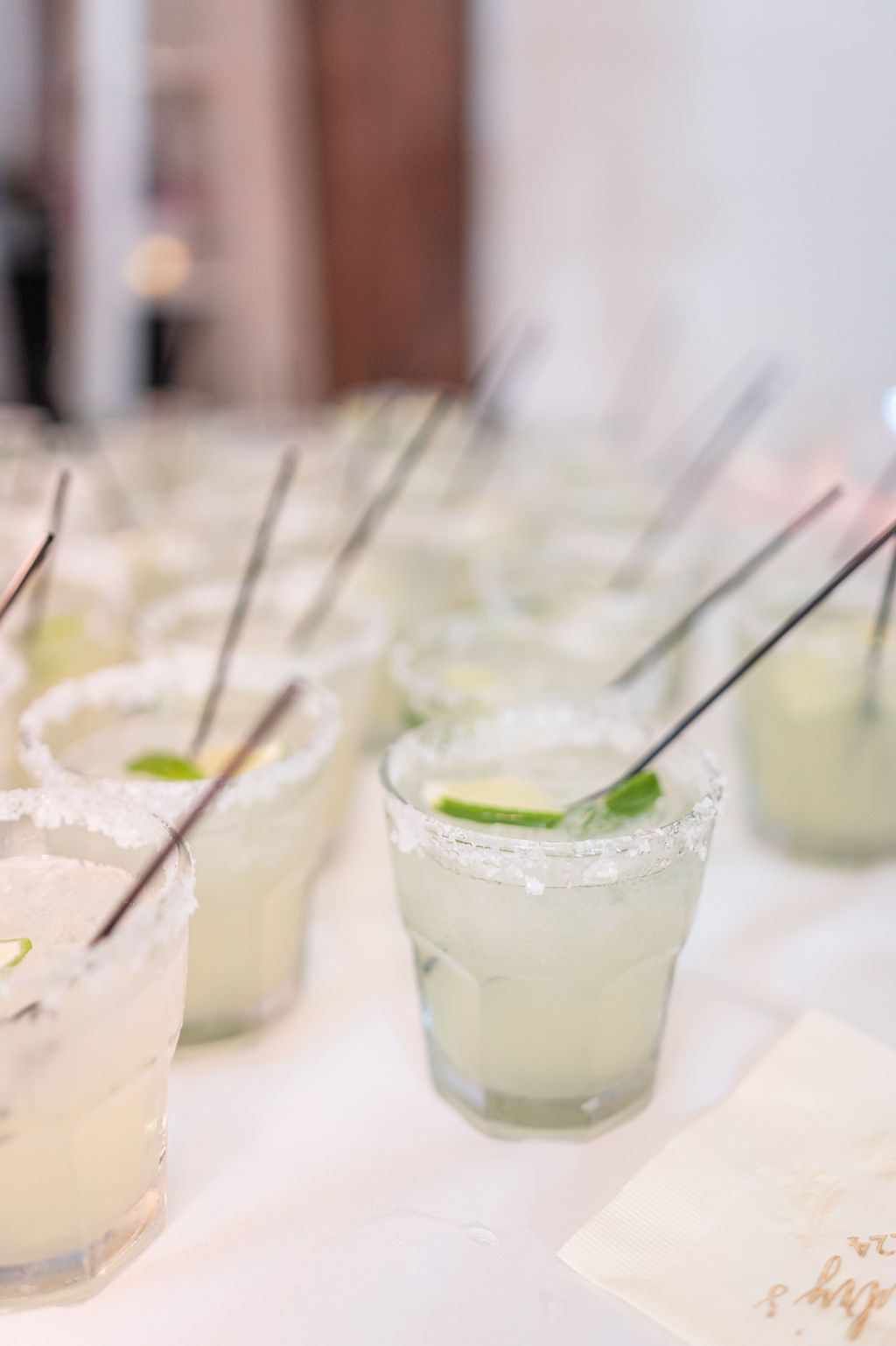 Margaritas in glasses with salt rims and lime wedges, arranged on a white table with straws.