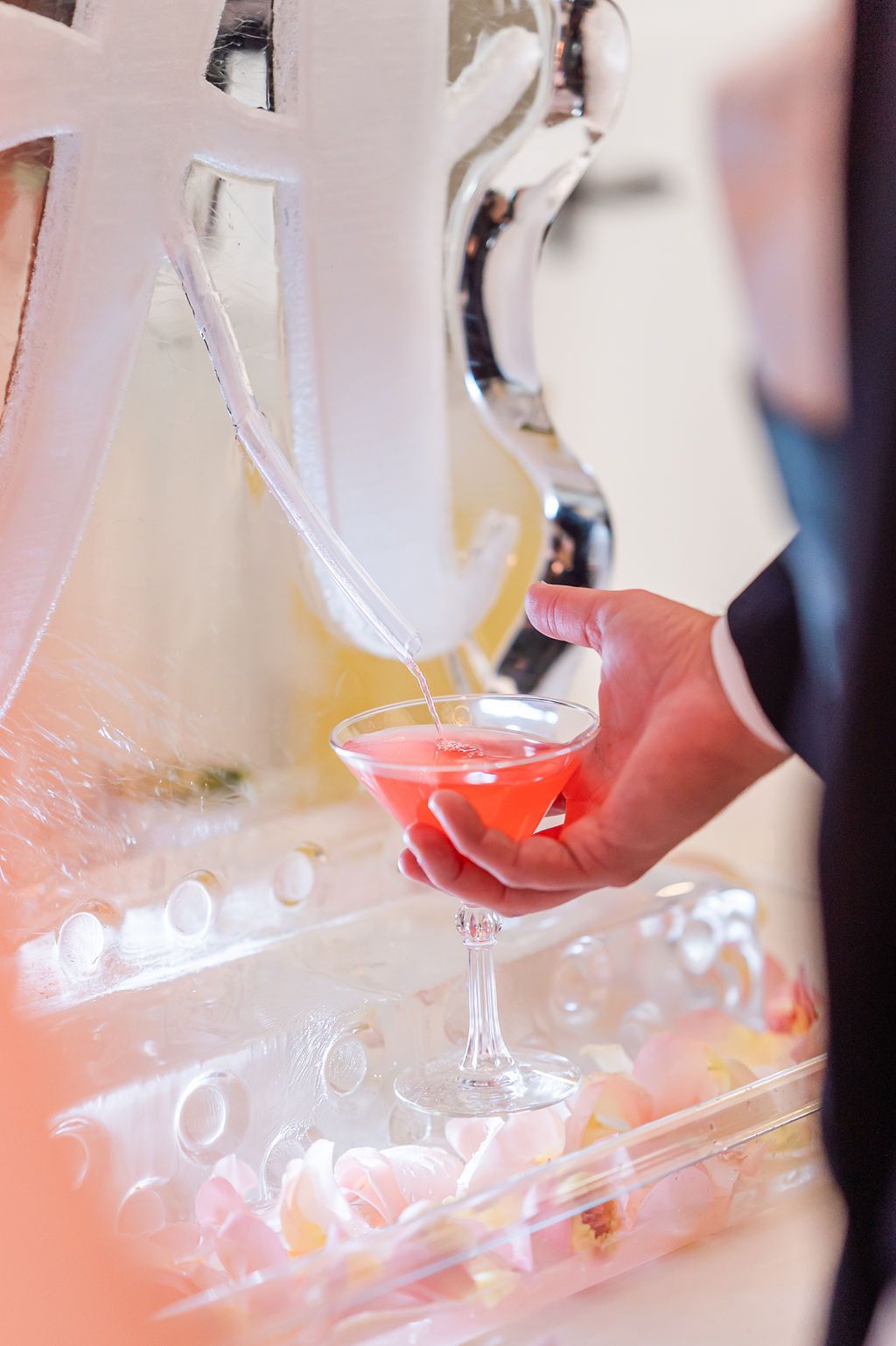 A hand pours pink liquid into a martini glass from an elaborate ice sculpture at an event.