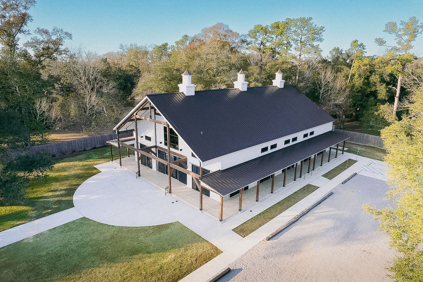 White barn with black roof, wooden accents, and a long porch, set on a lawn with trees in the background.