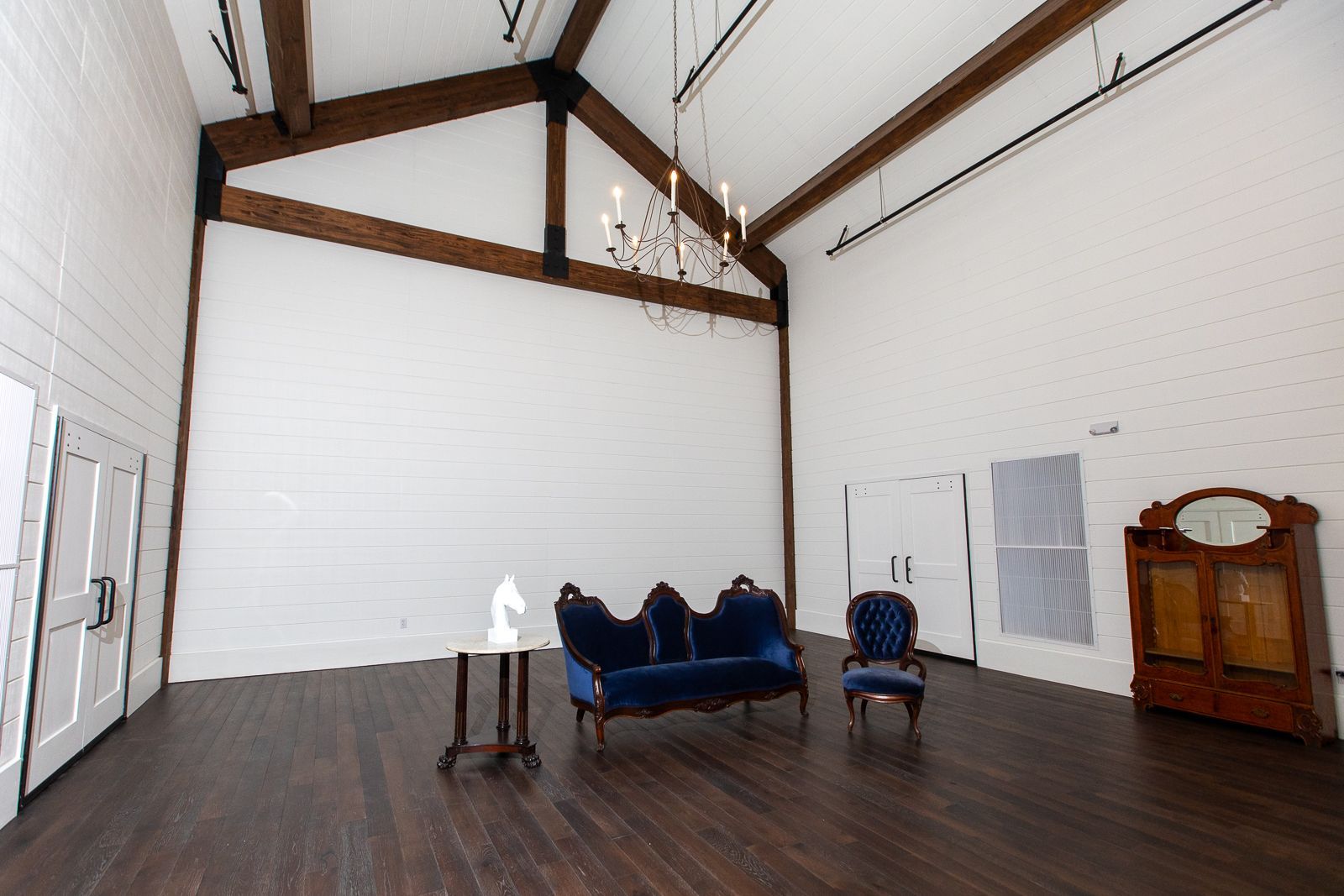 Empty room with exposed wooden beams, white walls, and dark wood floor. Blue velvet furniture is in the center.