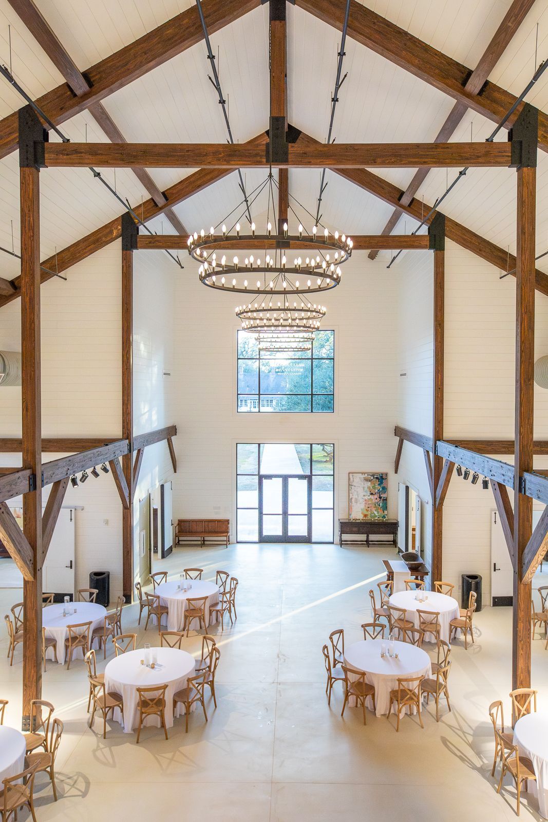 High-ceilinged barn interior with round tables, chandeliers, and wooden beams.