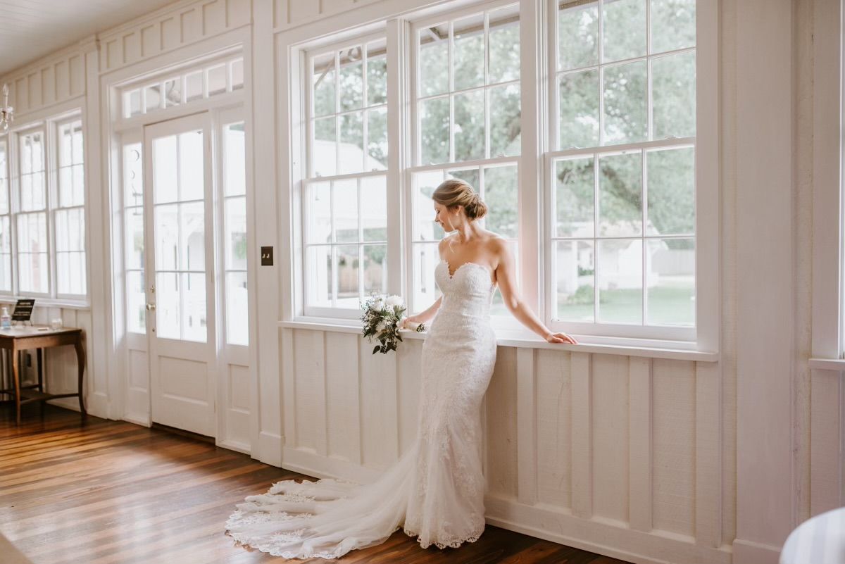 Bride in a white gown leans on a windowsill, holding a bouquet, looking out a large window.