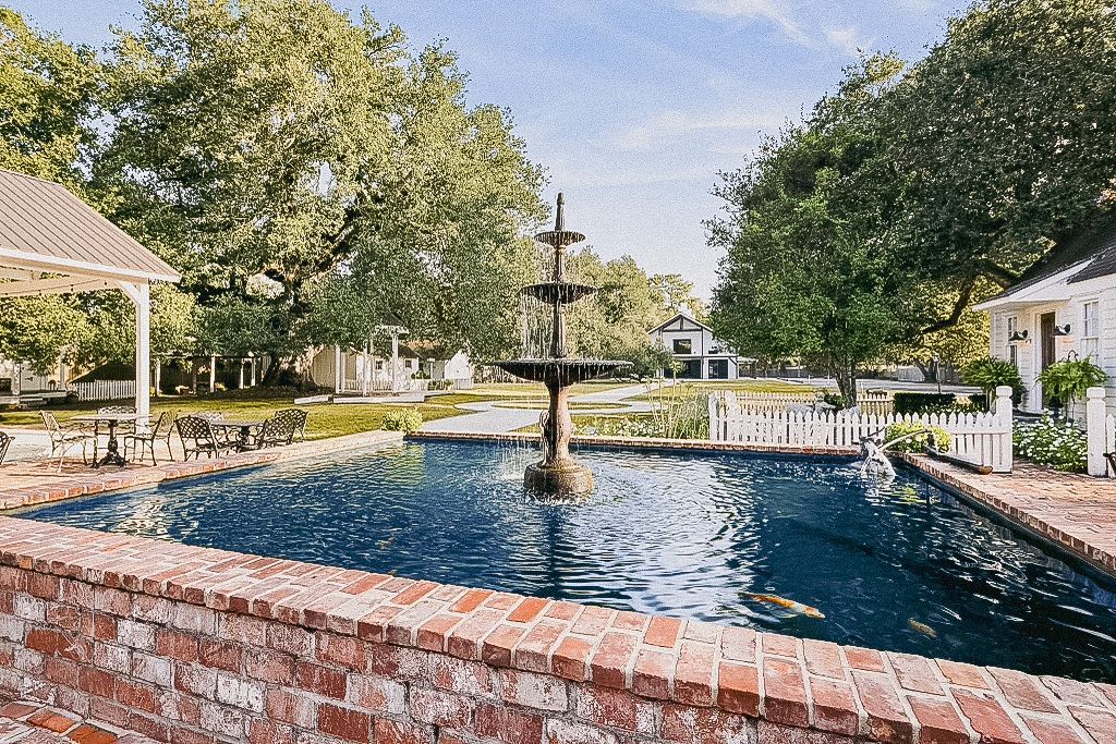 Brick-lined pond with fountain, white picket fence, and trees, creating a peaceful outdoor setting.