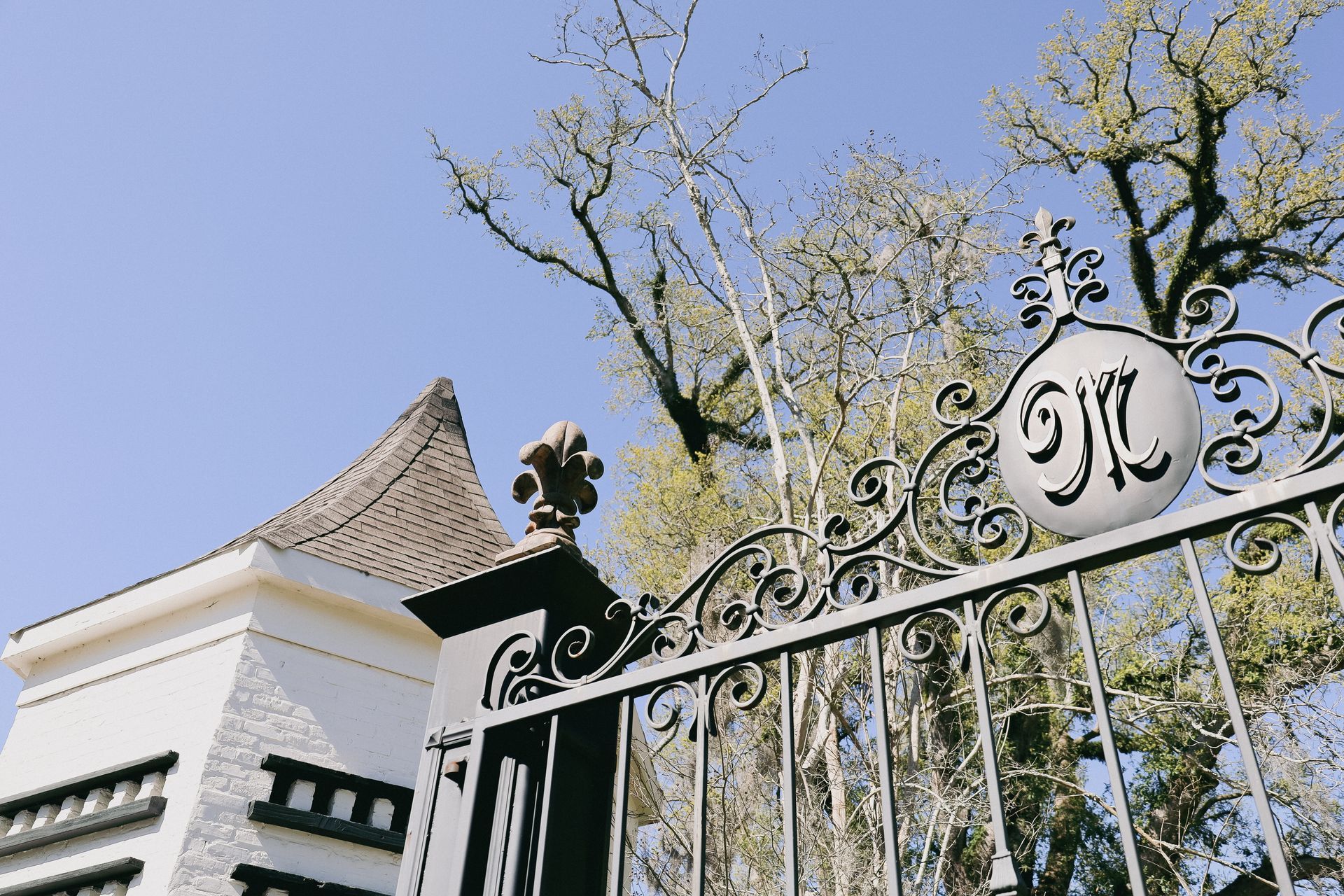 Ornate wrought iron gate with initials, adjacent to a white building with a brown roof, against a blue sky.