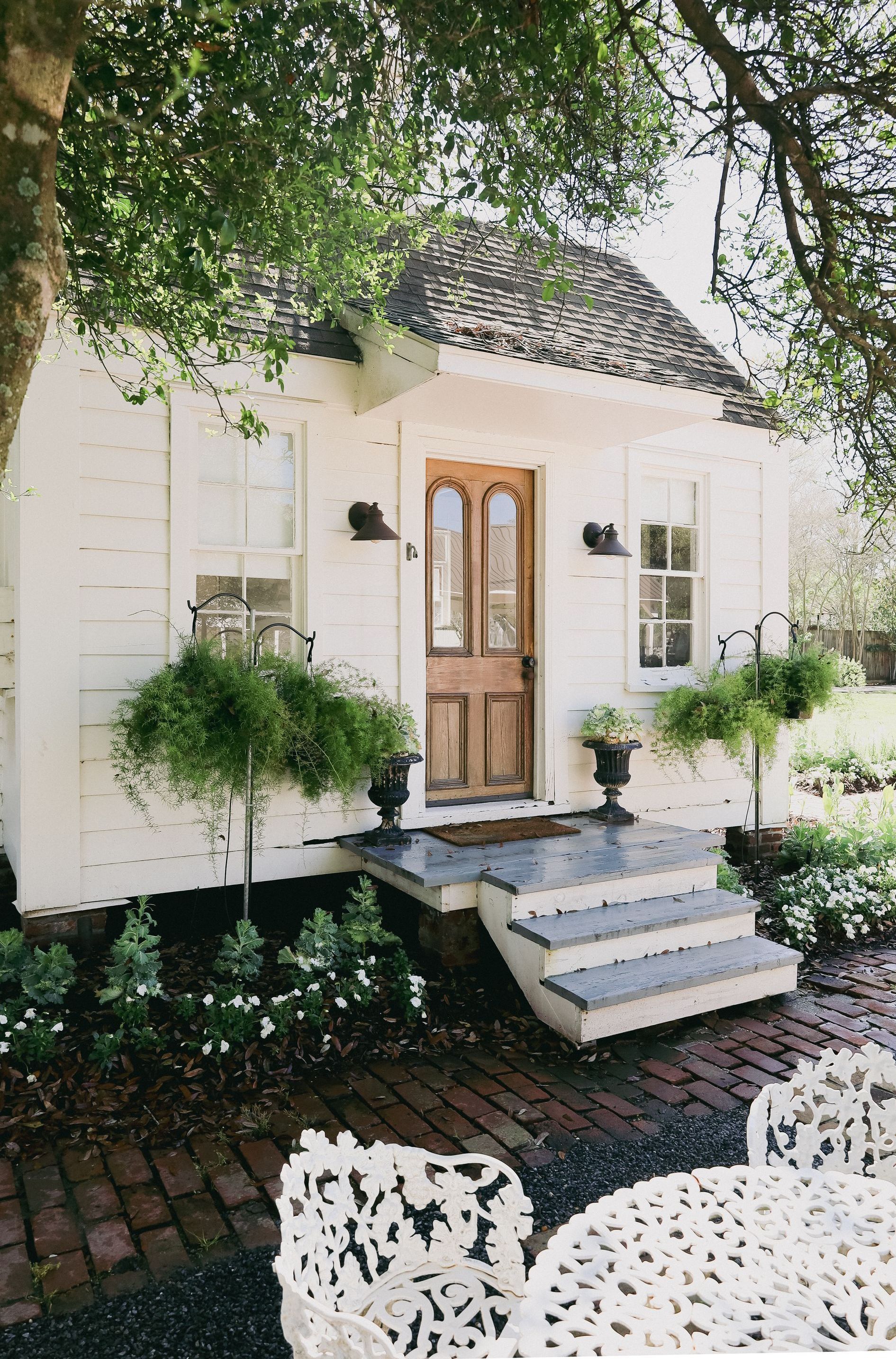 White cottage with brown door and arched roof, brick pathway, ornate outdoor table and chairs.