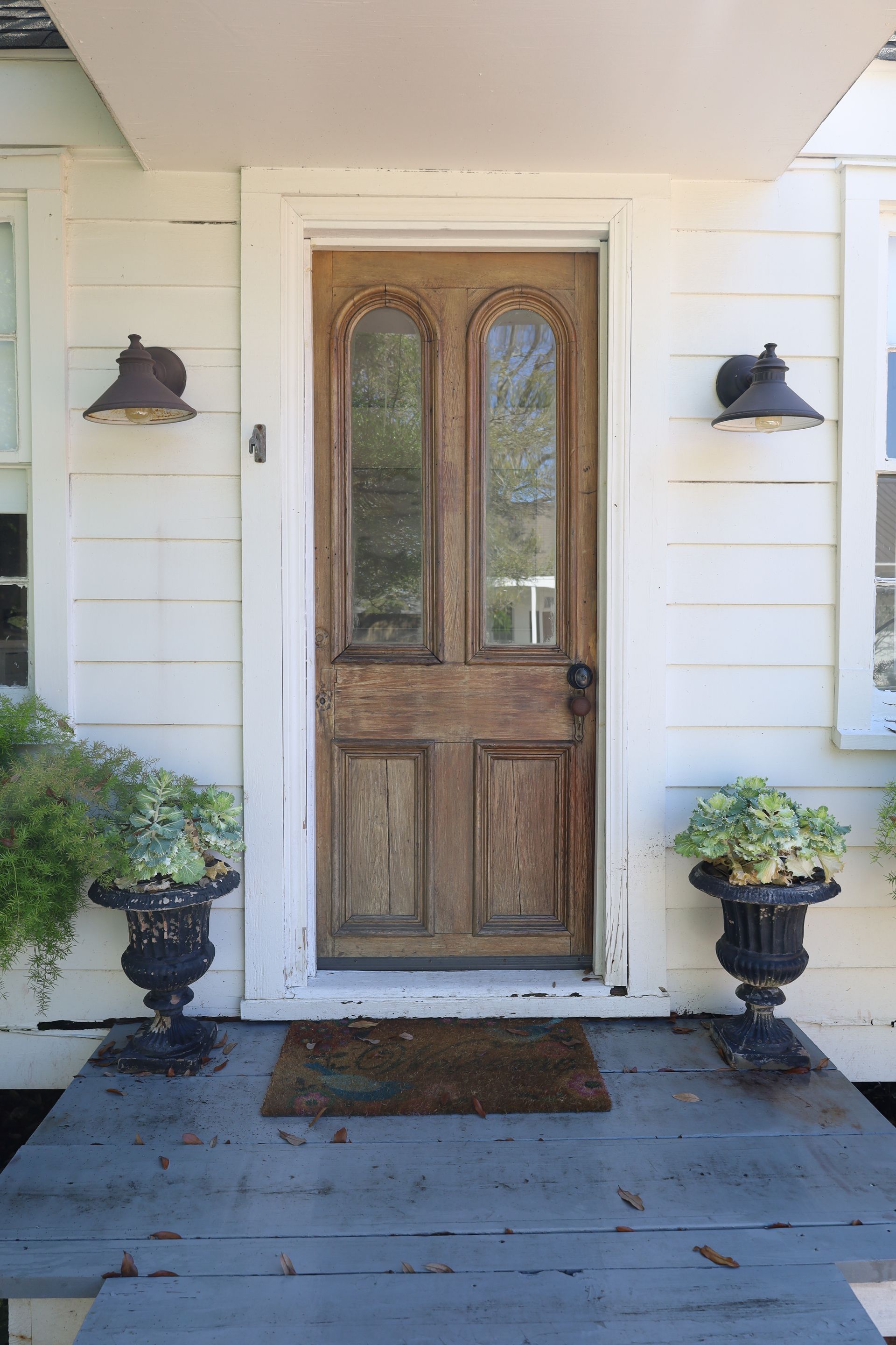 Weathered wooden front door with glass panels, flanked by sconces and potted plants.