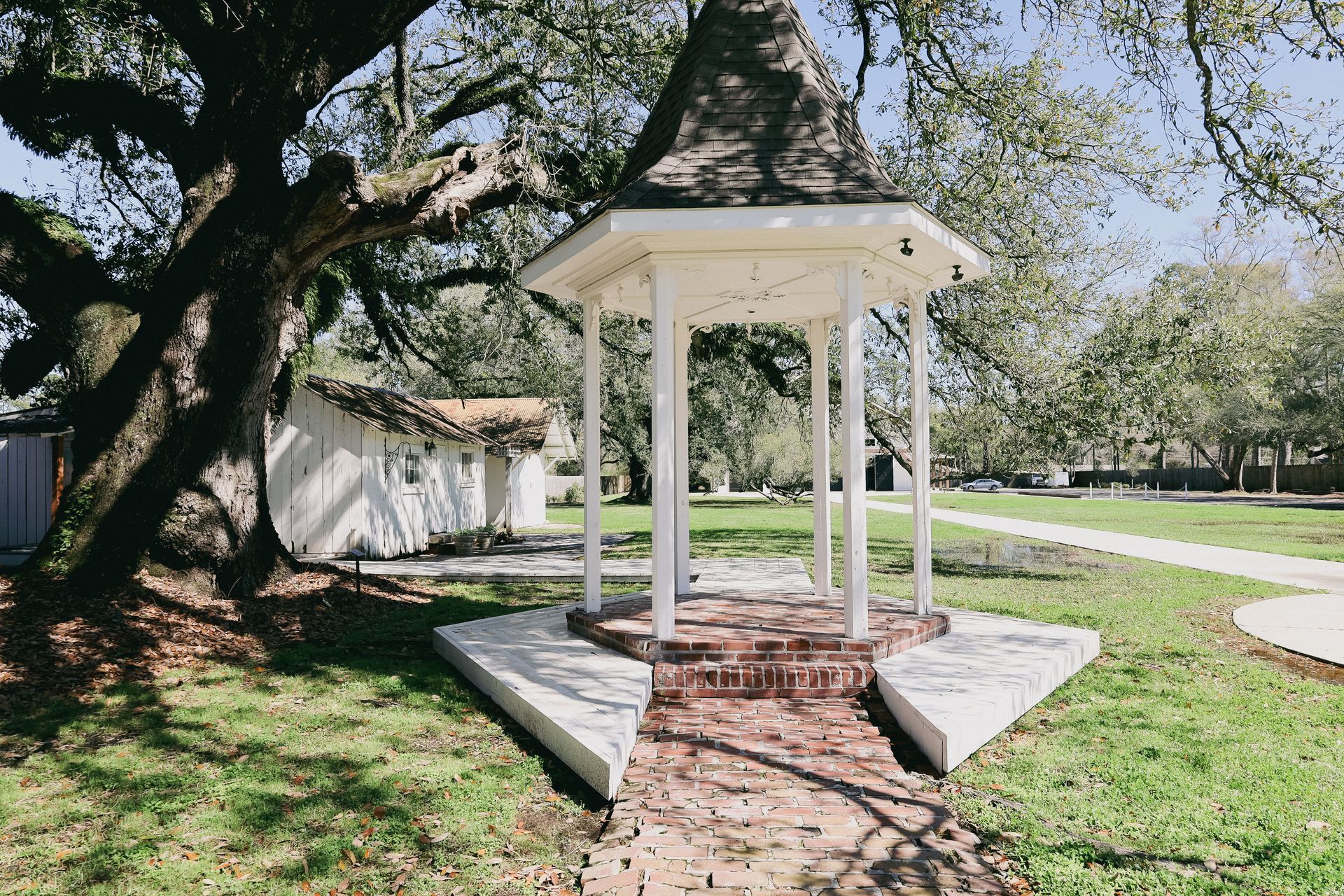 White gazebo with brick floor in a grassy park, a building and large tree in background.