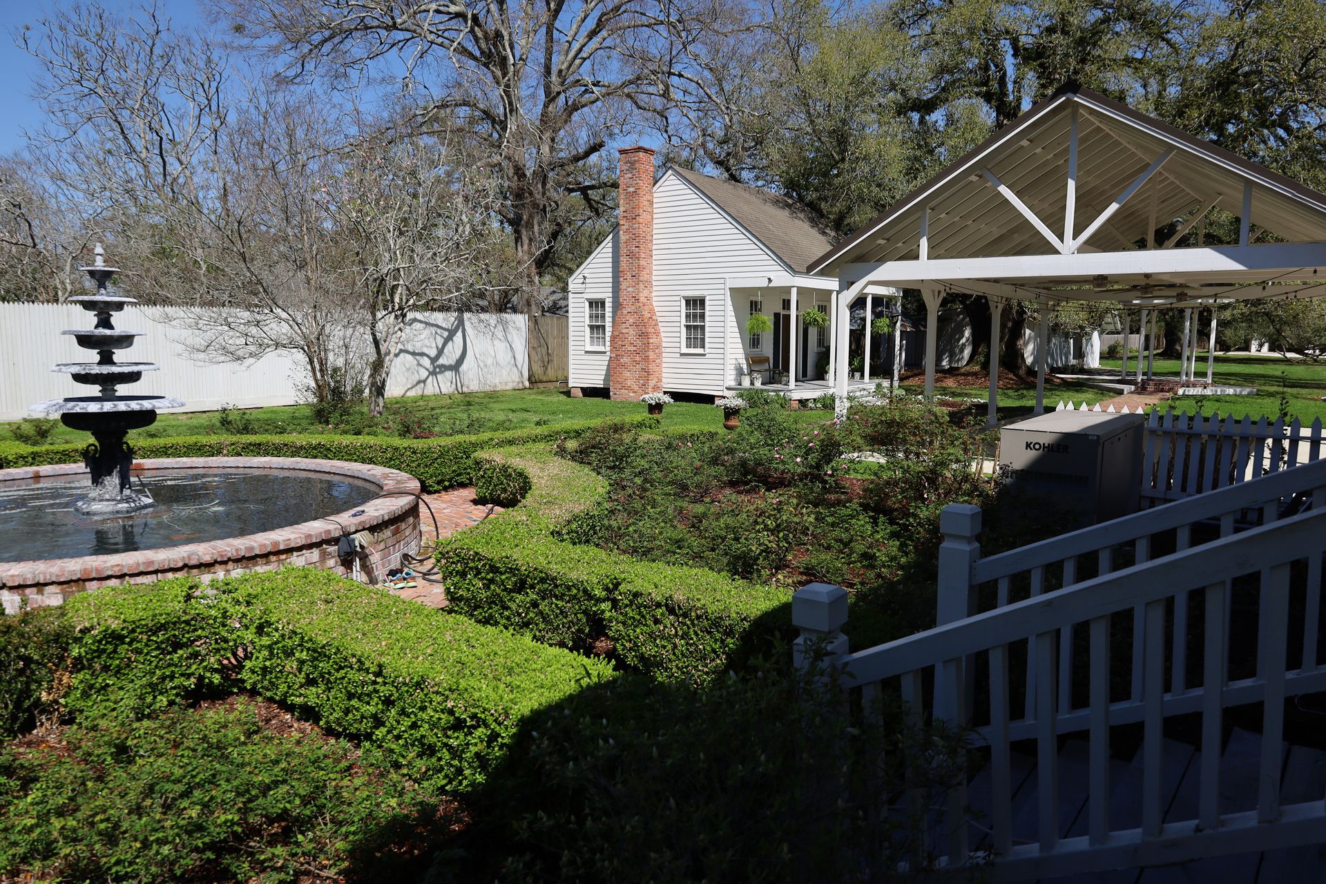 White building and gazebo with brick chimney in a garden with a fountain and manicured hedges.