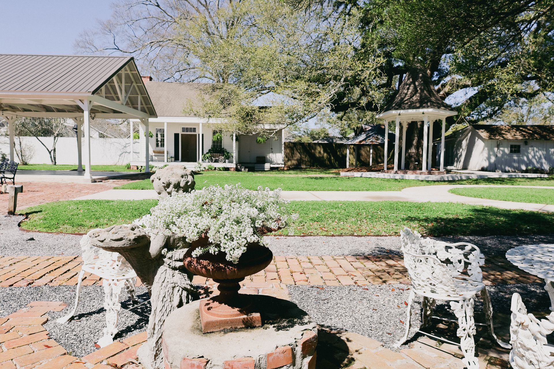 Lush garden with brick paths, white-columned structures, and a house in the background.