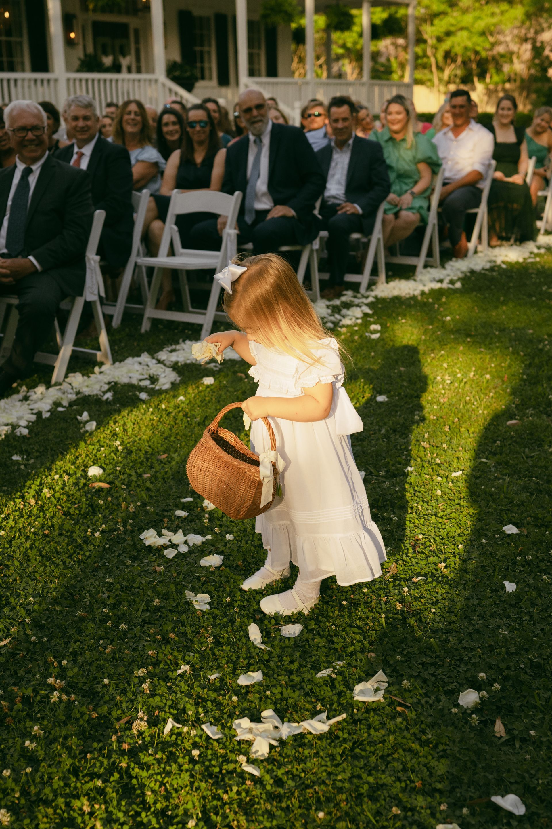 Flower girl scattering petals at outdoor wedding. Guests watch from white chairs.