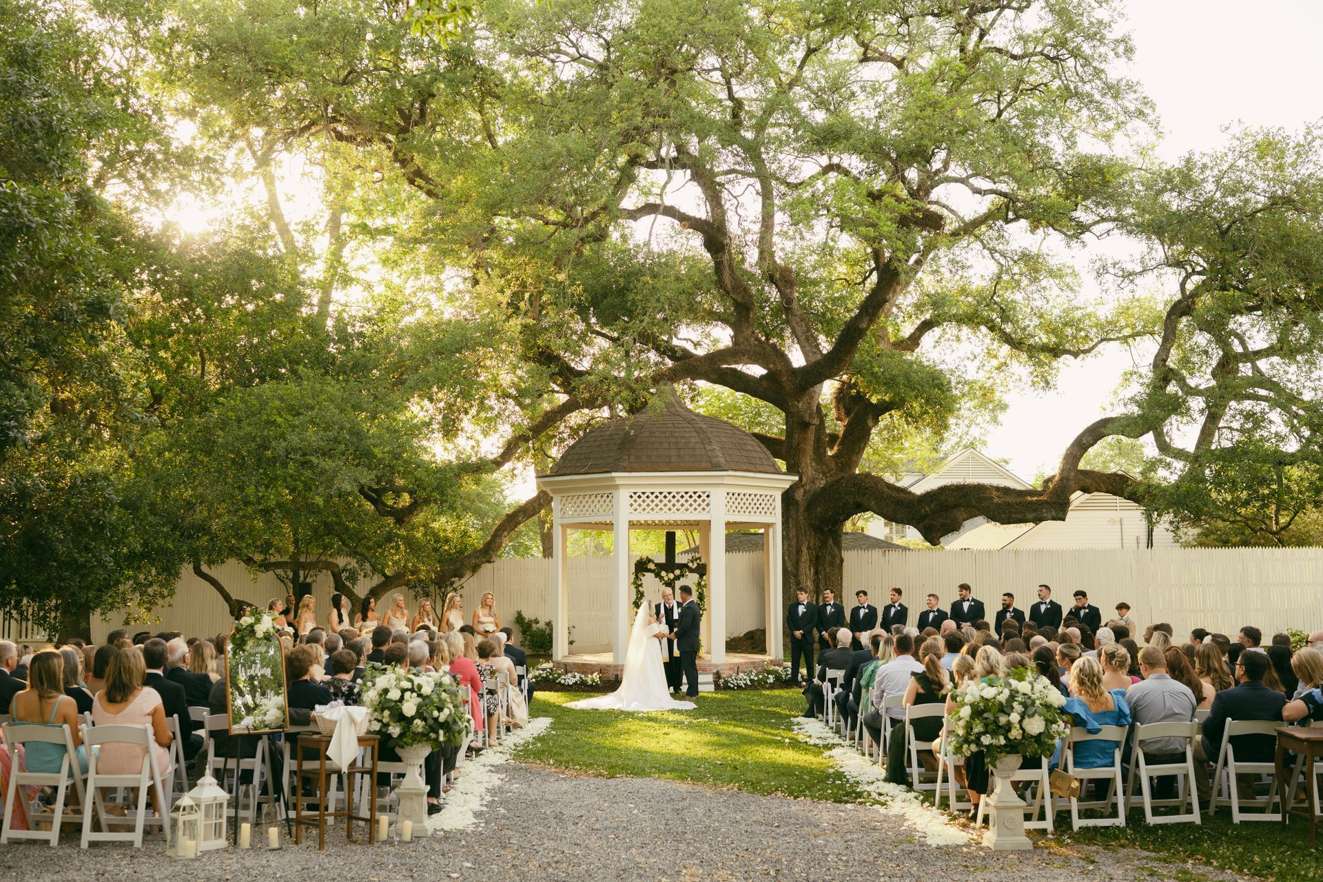 Wedding ceremony under a large tree with a gazebo. Guests seated, couple at the altar.