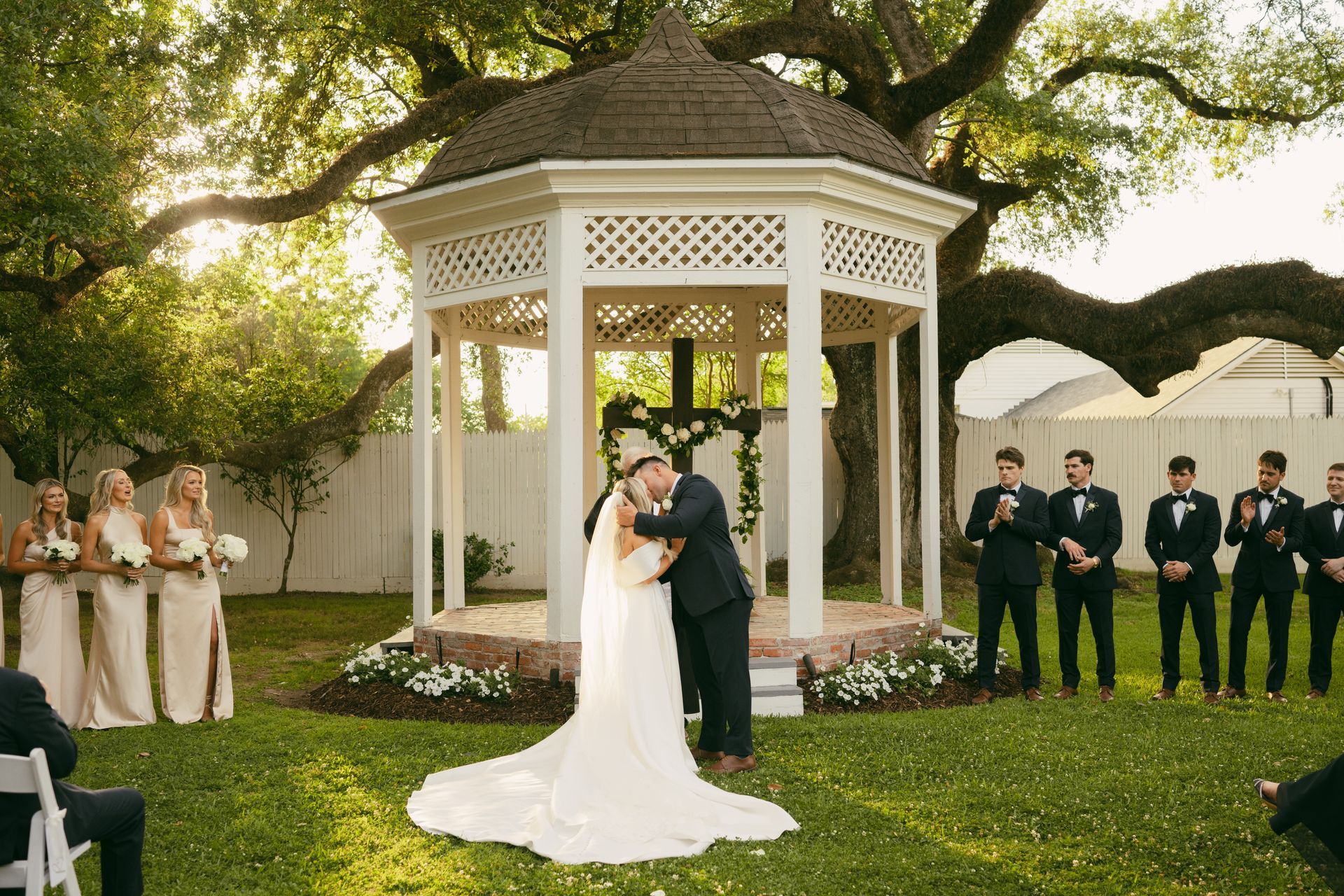 Bride and groom kissing under a gazebo during outdoor wedding ceremony. Groomsmen, bridesmaids watch.