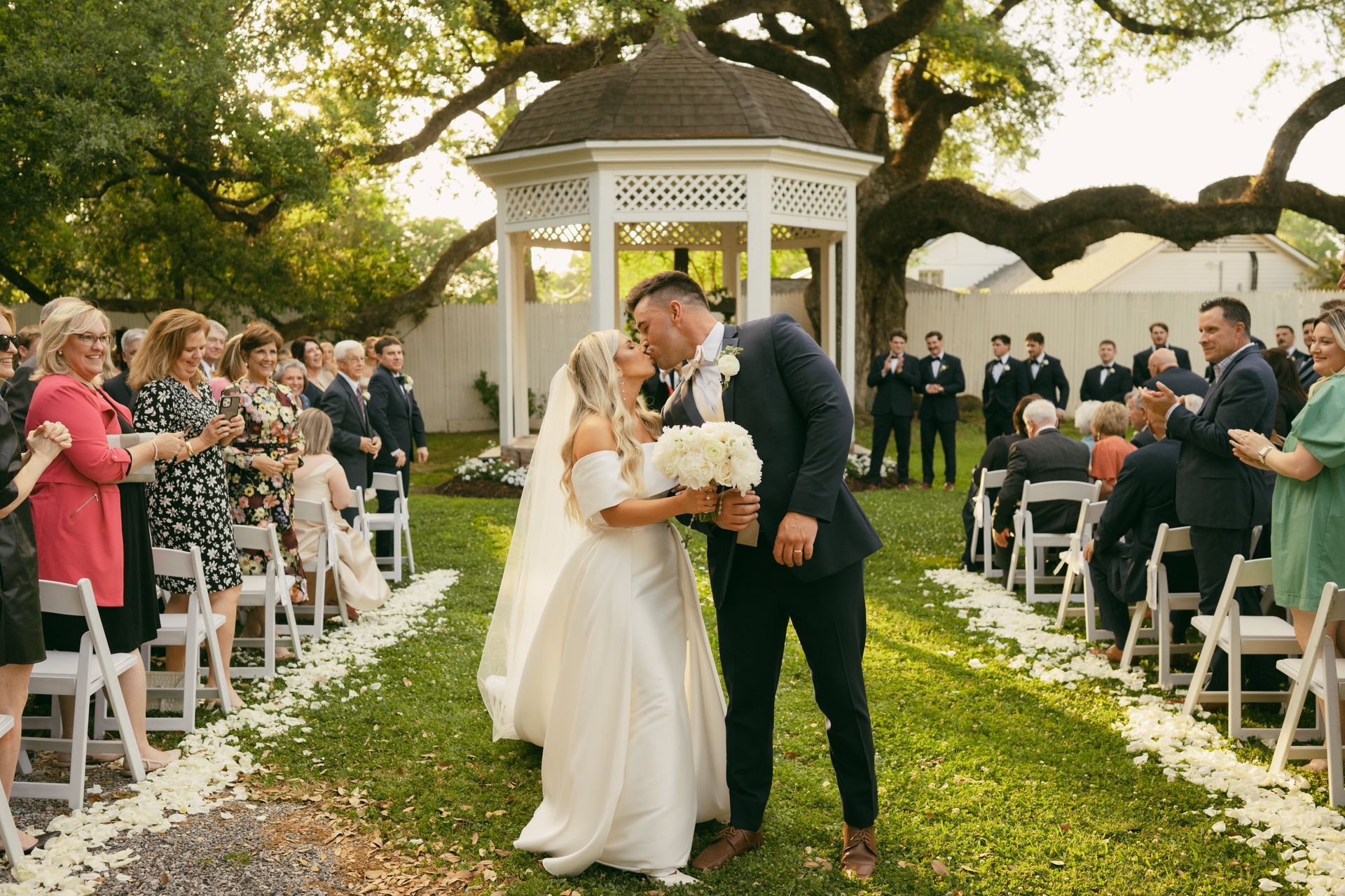 Newlyweds kiss in a garden lined with guests and white chairs, under a gazebo.