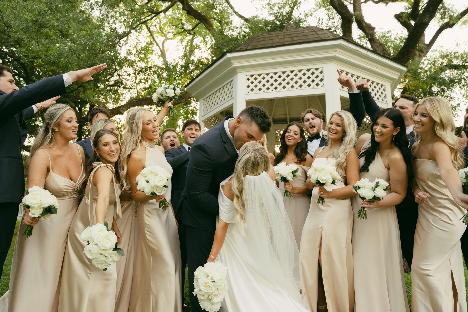 Wedding group photo: Bride and groom kissing, surrounded by bridesmaids and groomsmen near a gazebo, everyone celebrating.