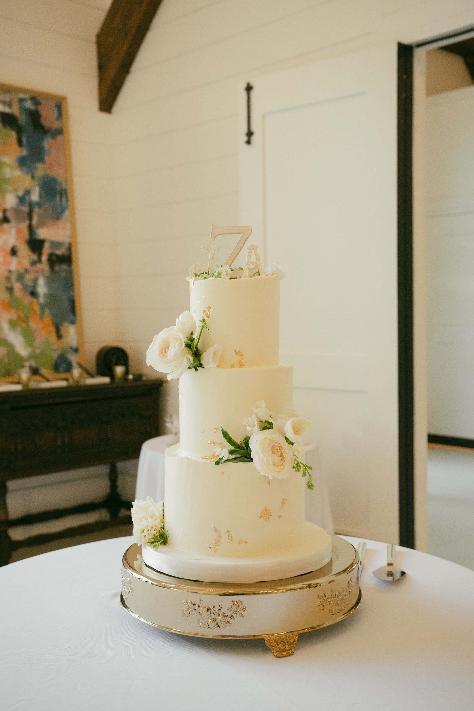 Four-tiered wedding cake, off-white frosting, floral accents, on a decorative gold stand, set on a white tablecloth.
