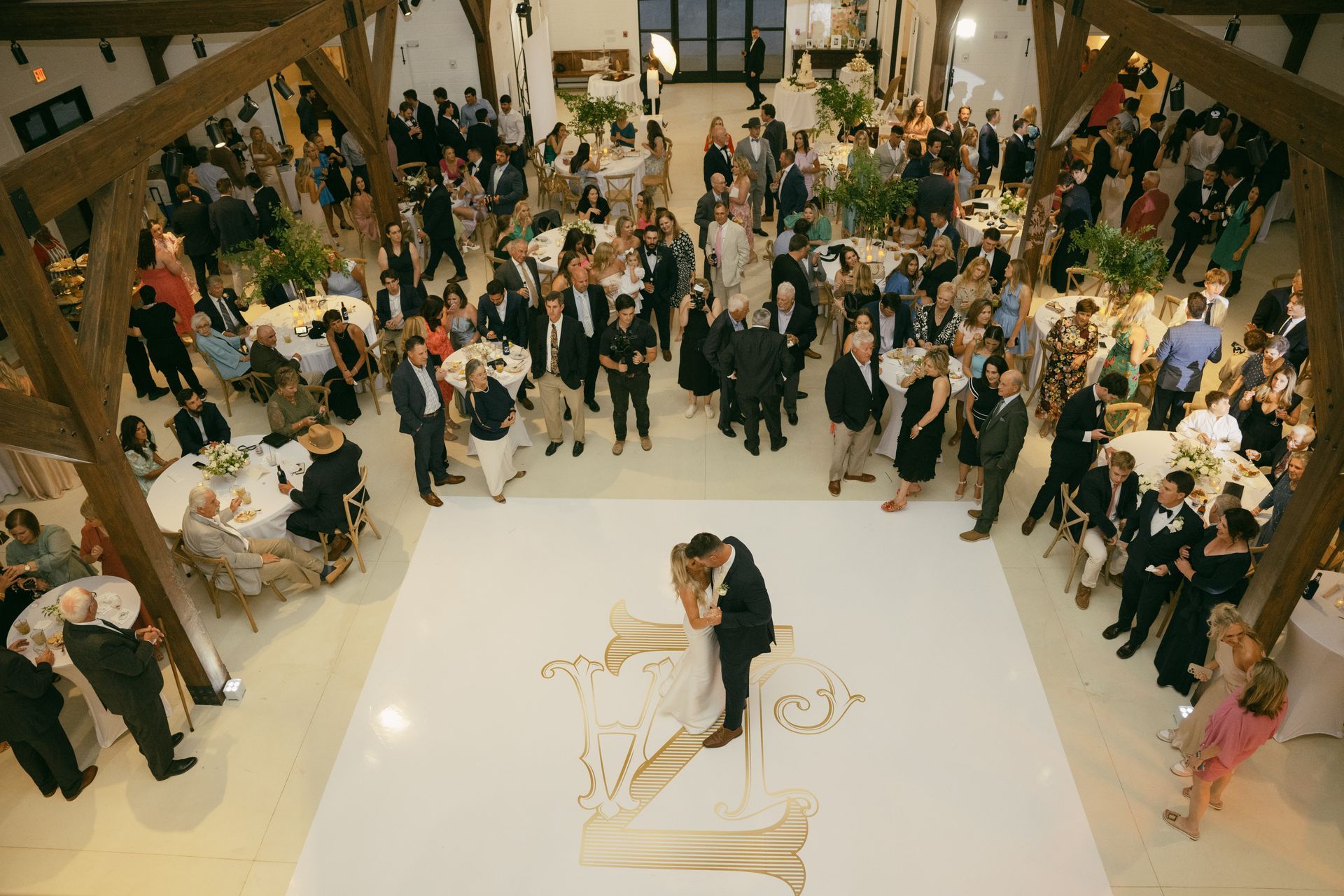 Wedding reception: couple dances on a decorated floor as guests watch in a large hall with wooden beams.