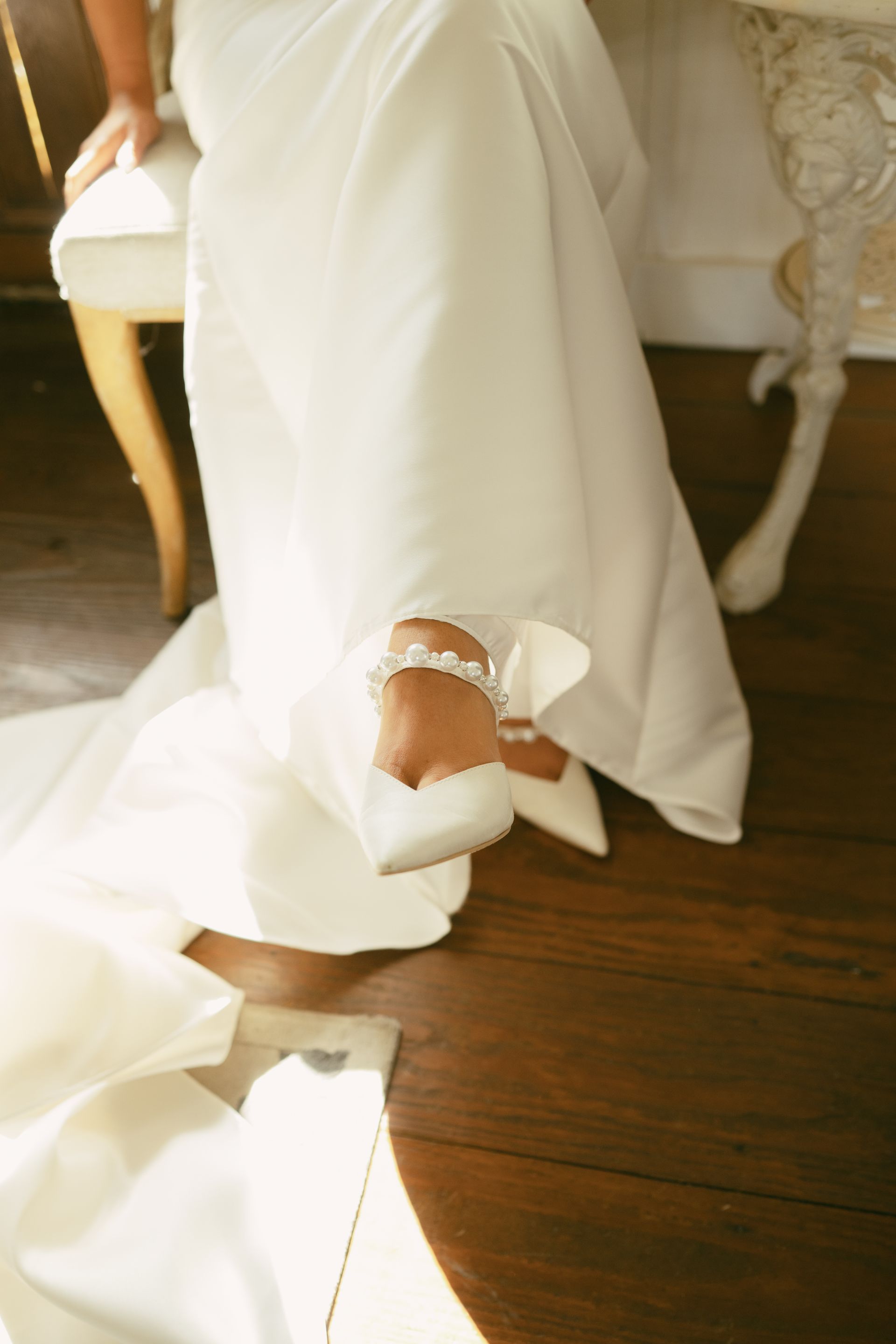 Bride in white gown and shoes, sitting; wooden floor, decorative chair.