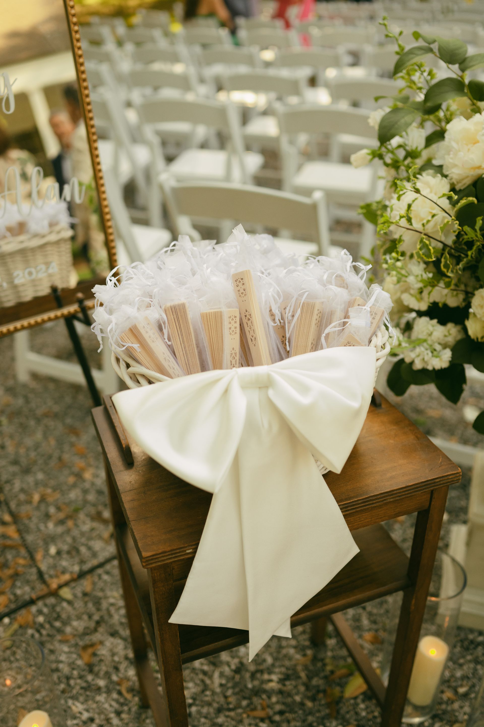 White basket of fans on a wooden chair, decorated with a large white bow, at an outdoor wedding.