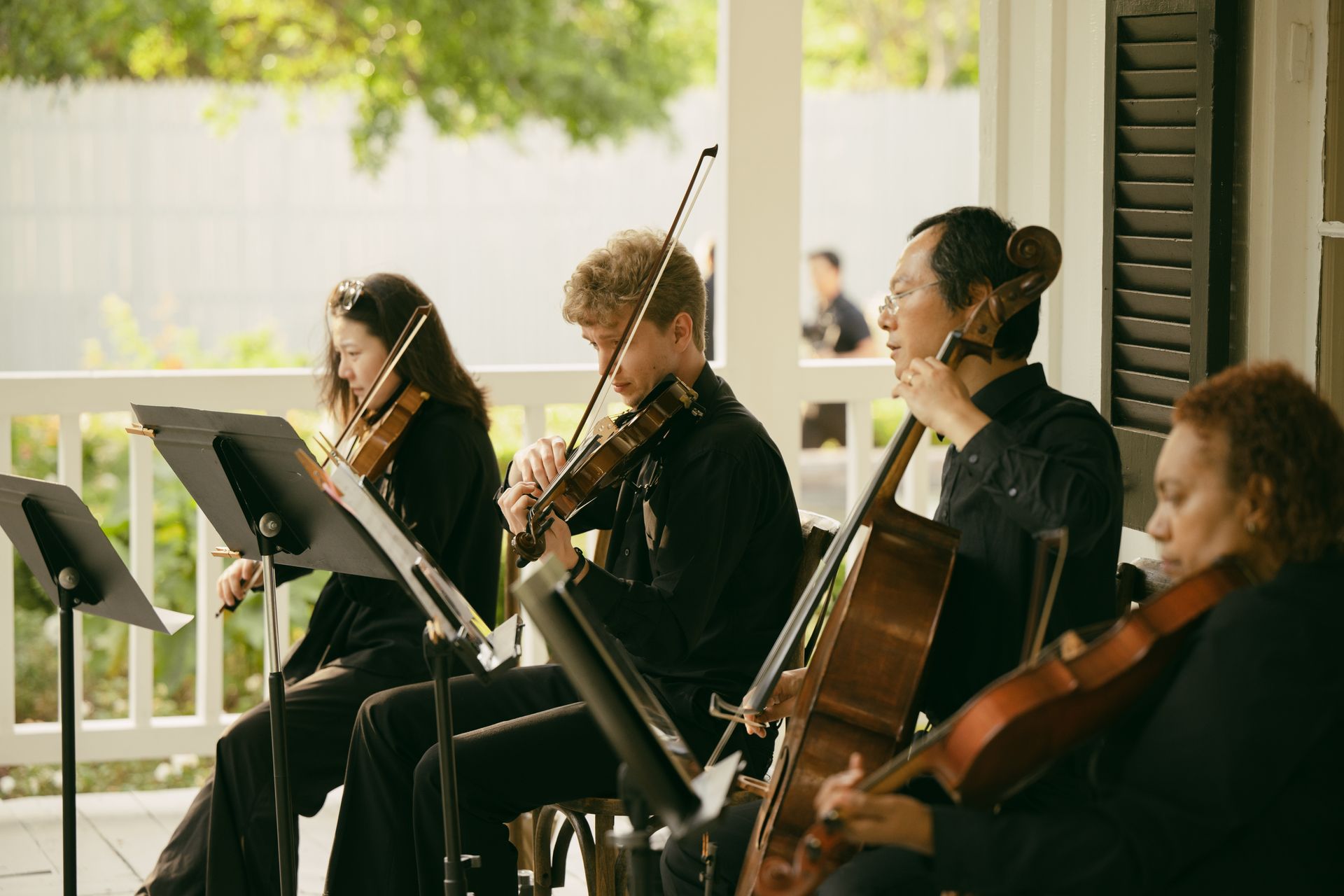 String quartet performing on a porch. Musicians play violins and cello, reading music. Sunlight.