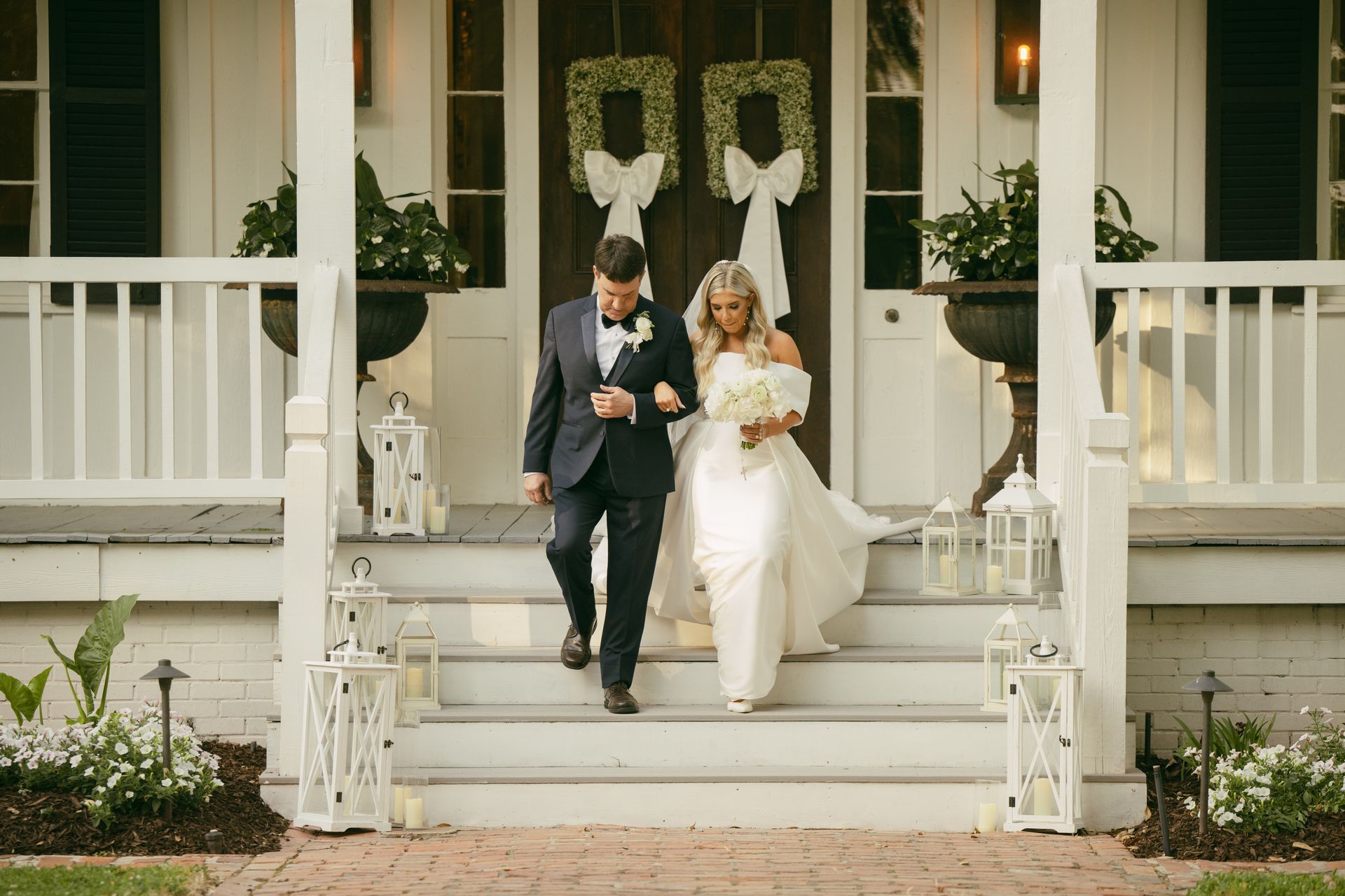 Bride and groom descend white porch steps after ceremony, floral wreaths and lanterns in setting.