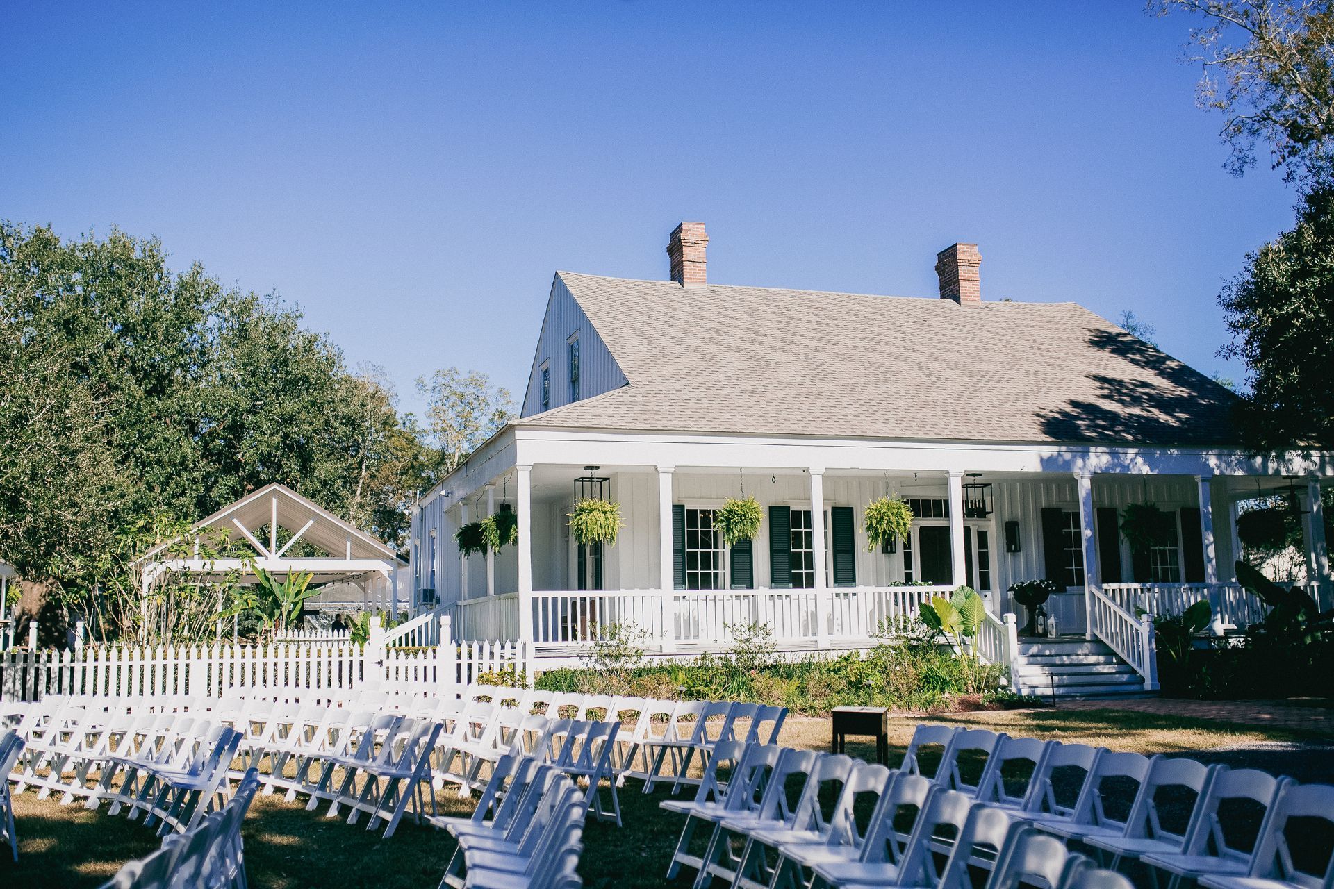 White house with a wrap-around porch, white fence, and rows of white chairs set up for an outdoor event.