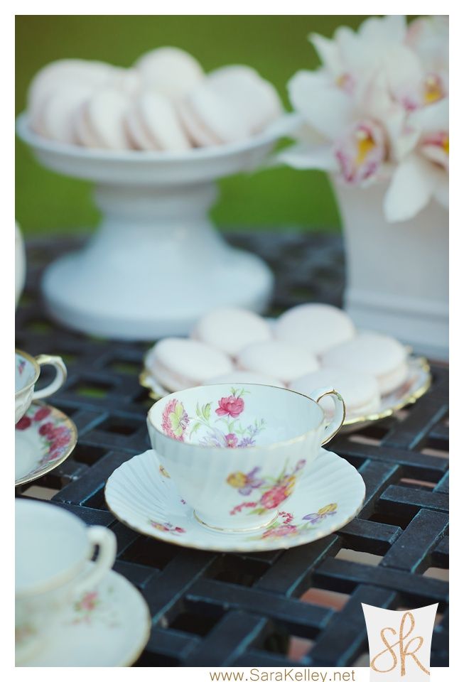 Tea cups with floral patterns on a table, sweets and flowers in the background.