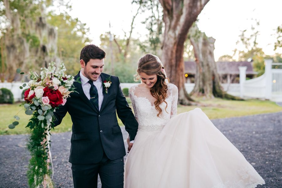 Newlyweds walk hand-in-hand, smiling. Groom in black suit, bride in white gown. Outdoor setting, holding bouquet.