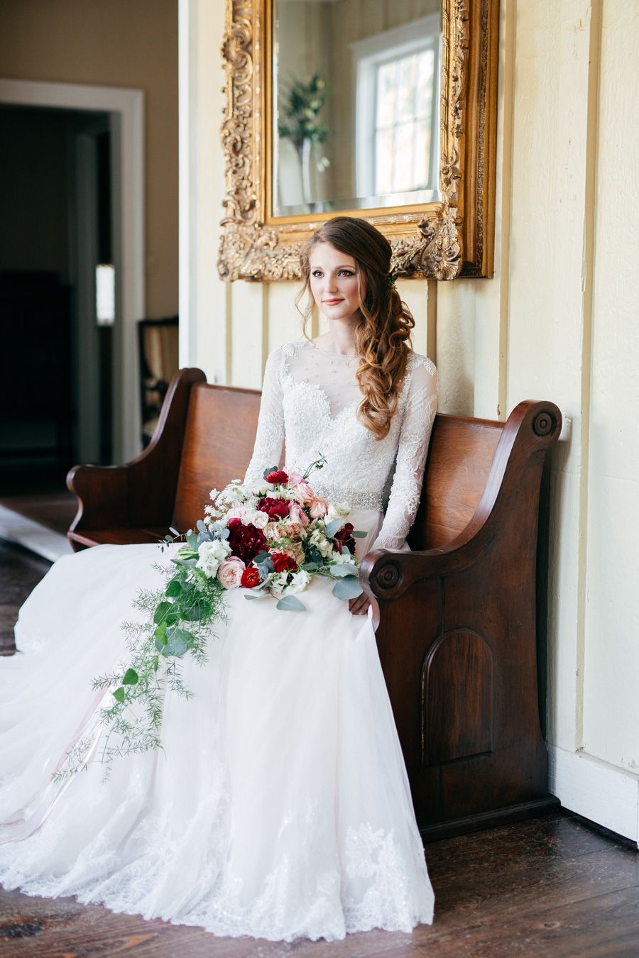 Bride in a white gown sits on a wooden bench, holding a cascading bouquet. She smiles in a hallway, near a mirror.