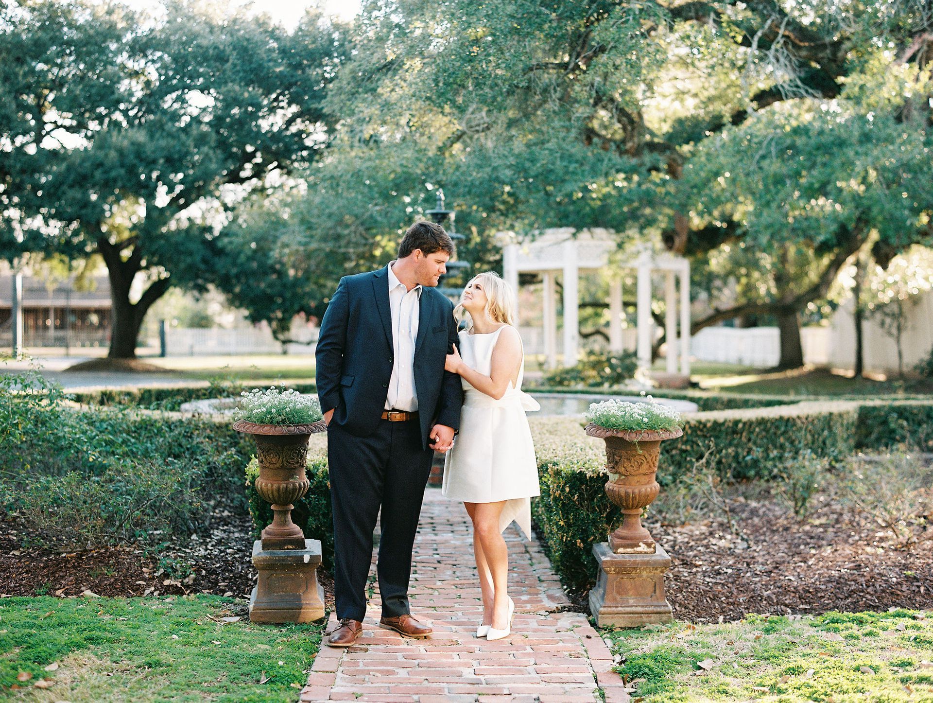 Couple walking hand-in-hand in a garden; man in a suit, woman in a white dress, gazing at each other.