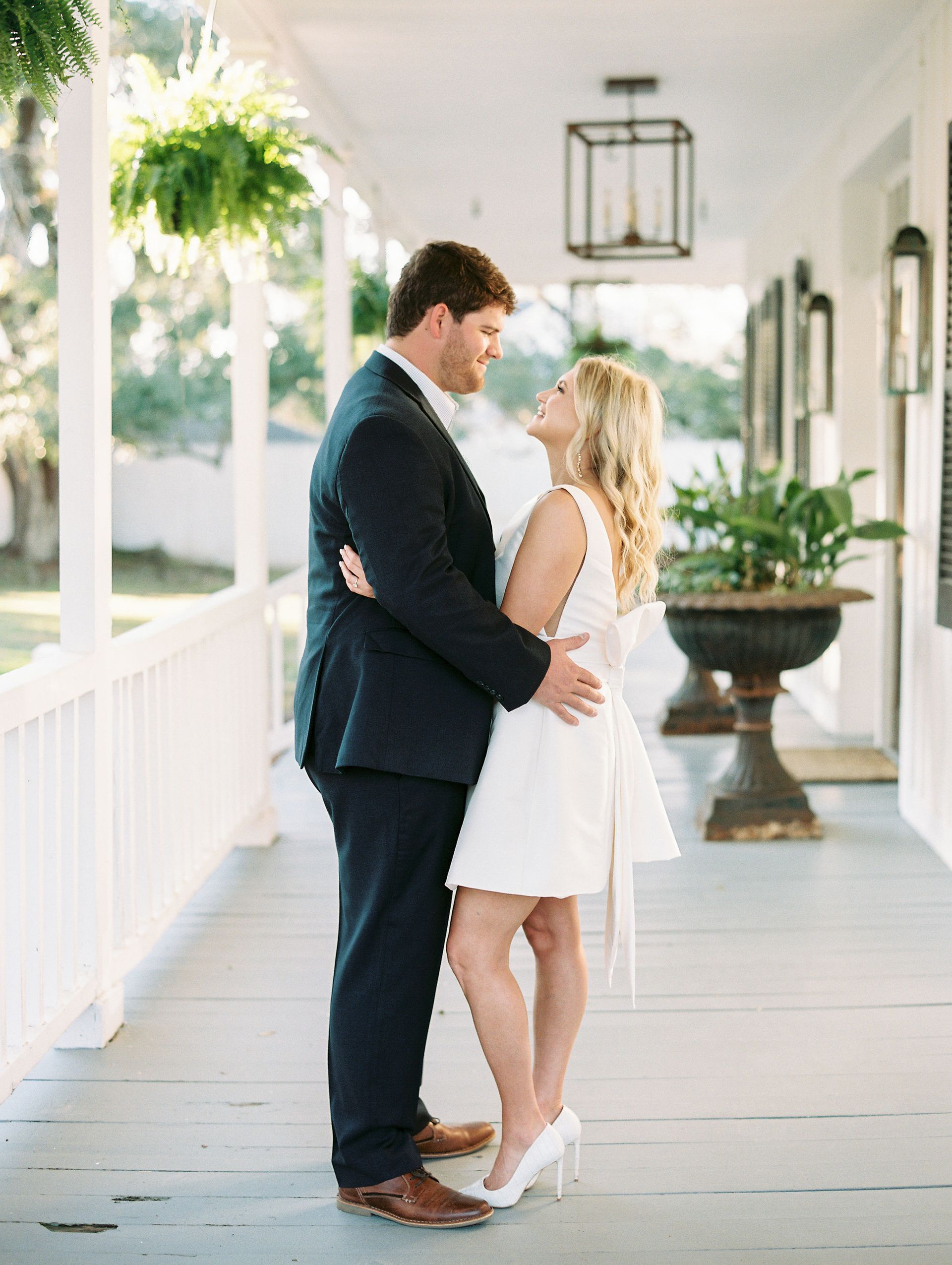 Couple embraces on a porch; the woman wears a white dress and heels, and the man wears a navy suit.