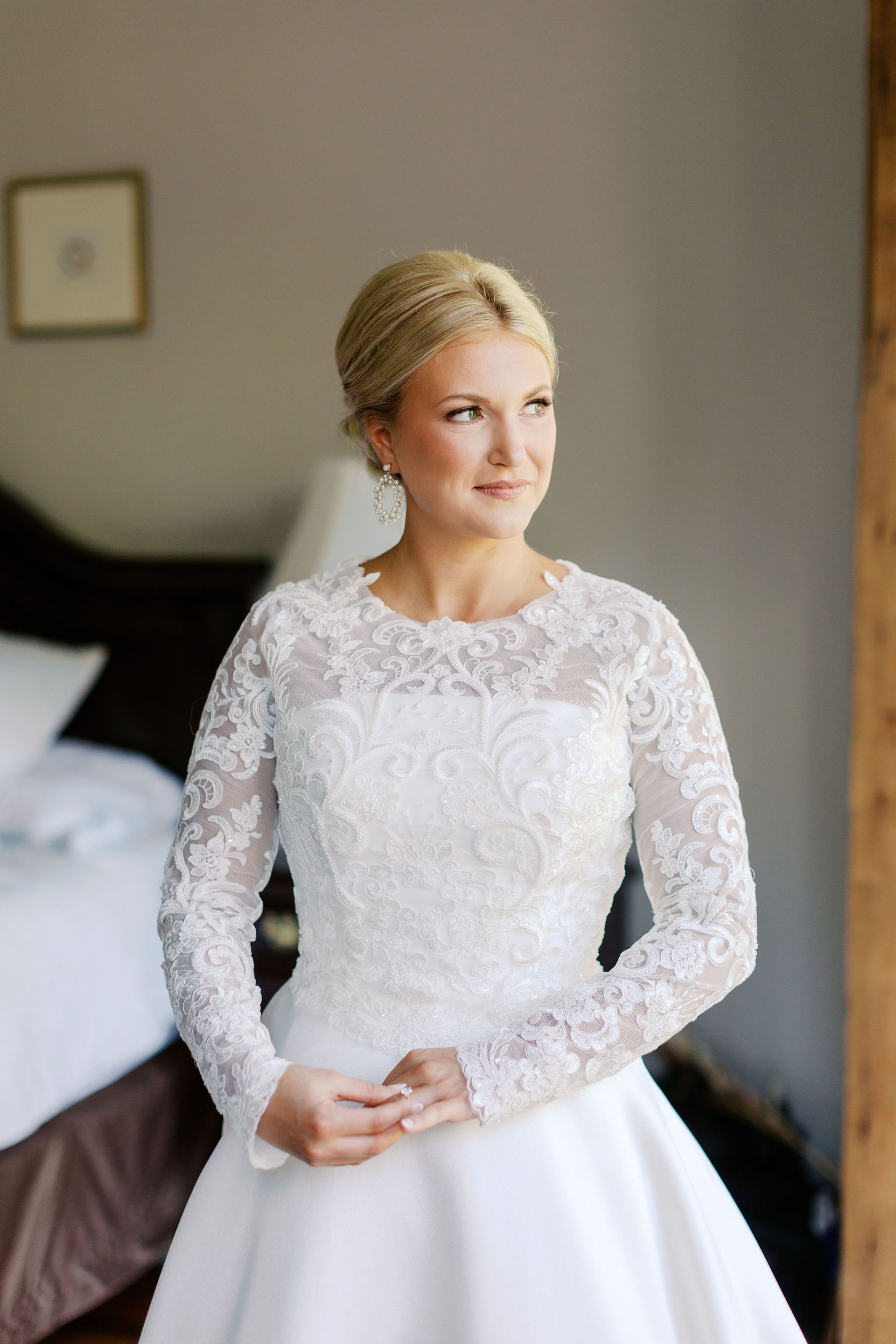 Bride in lace-sleeved white wedding dress, hands clasped, looking out the window.