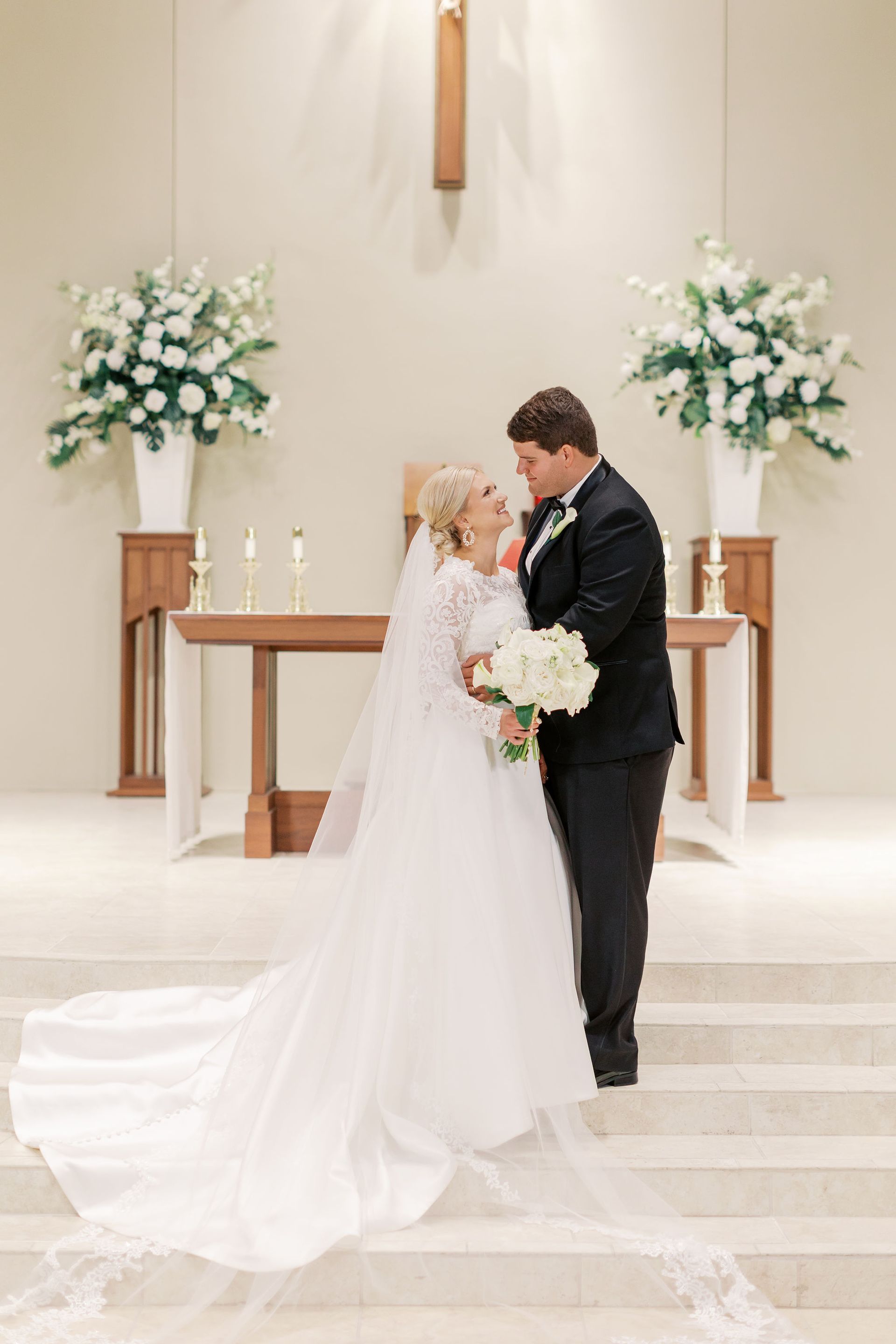 Bride and groom embrace in a church. She wears a white gown, he a tuxedo. White flowers adorn the altar.