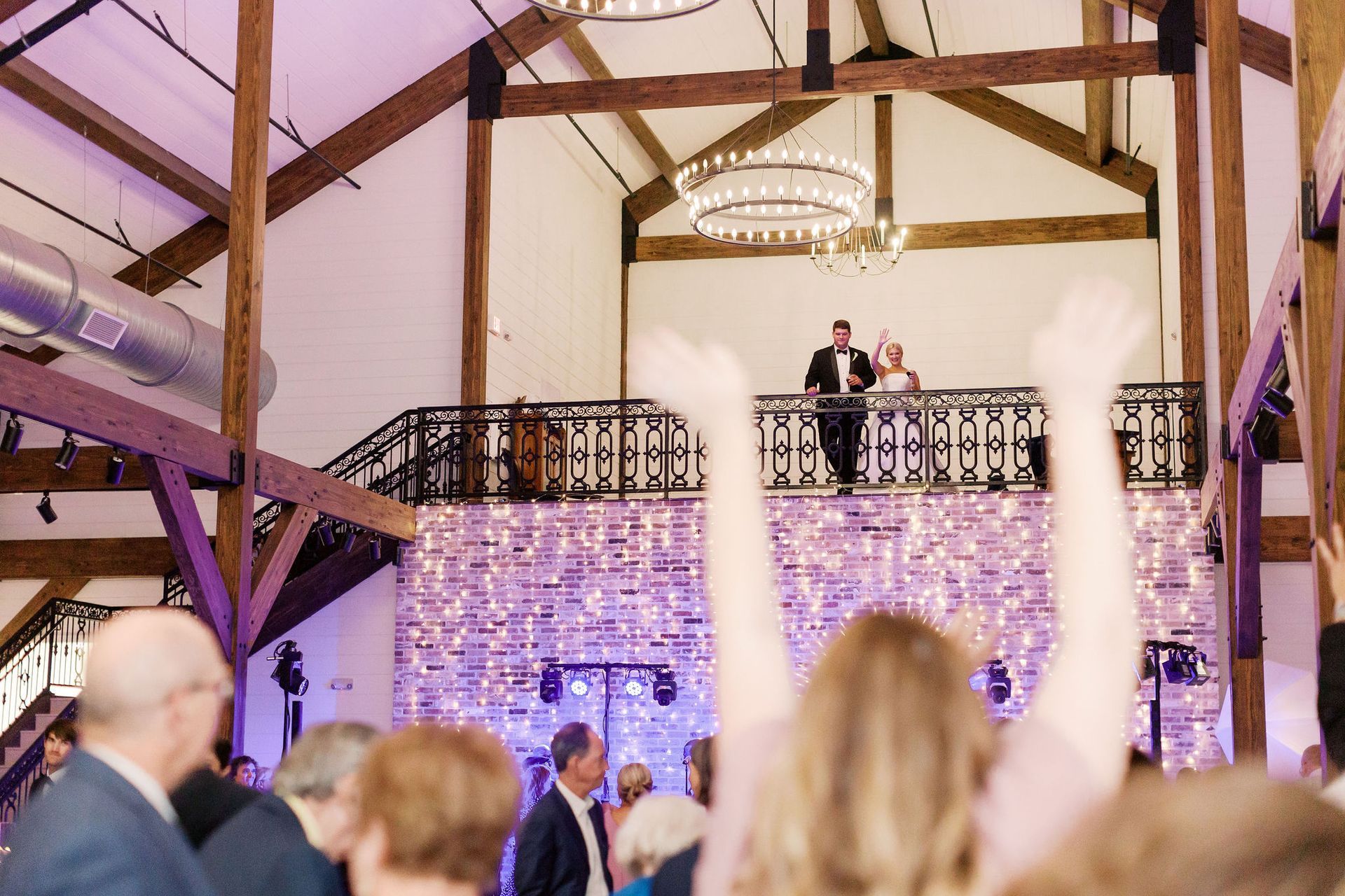 People celebrating below a balcony with couple, in a barn-like venue with string lights.