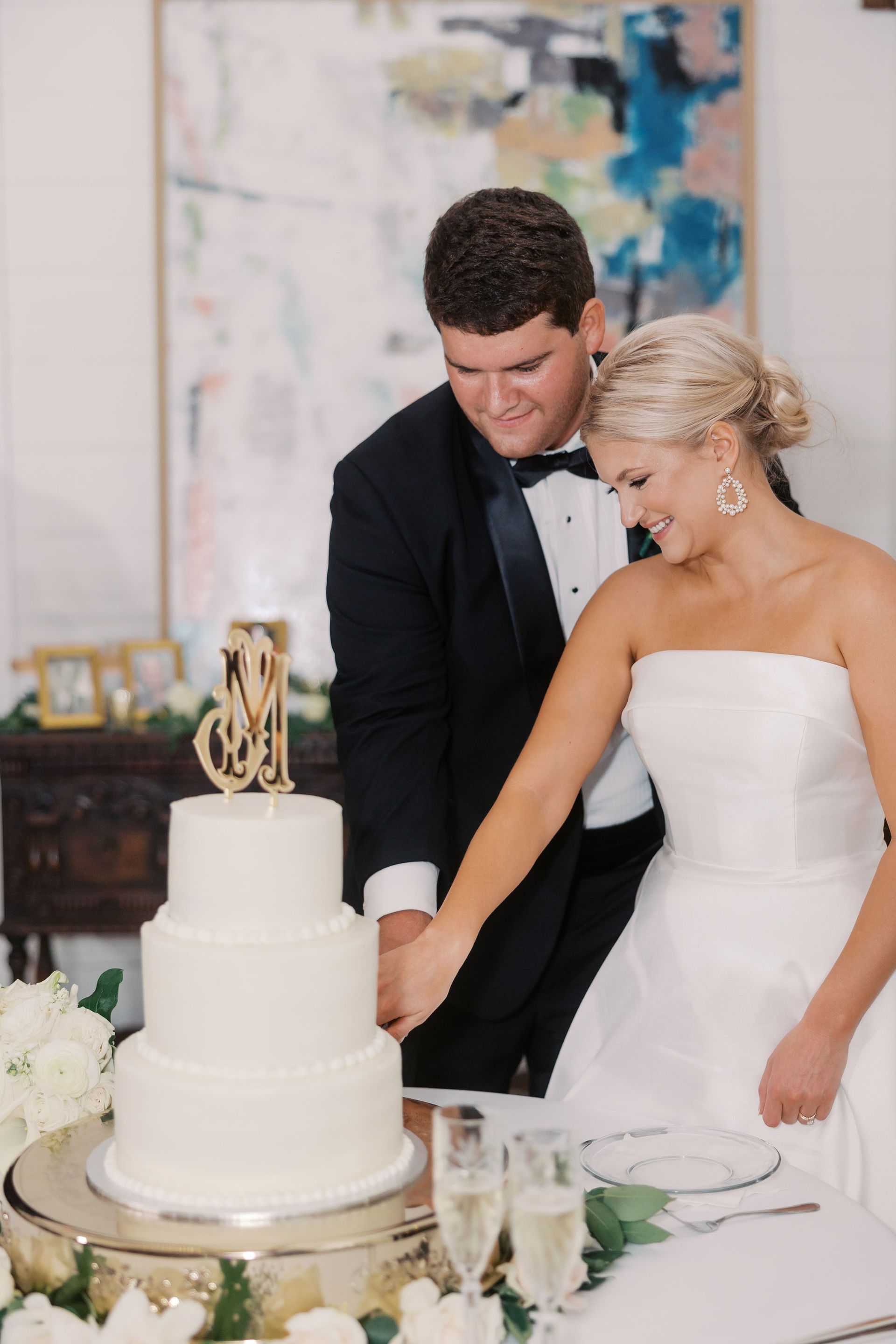 Couple in wedding attire cutting a three-tiered white cake with a monogram topper, smiling.