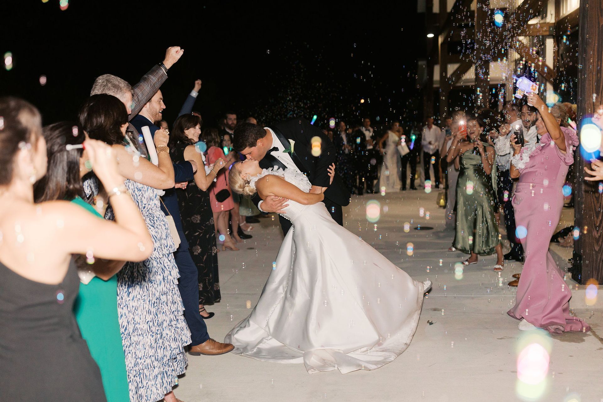 Newly married couple kisses under bubbles as guests cheer. Nighttime outdoor wedding.