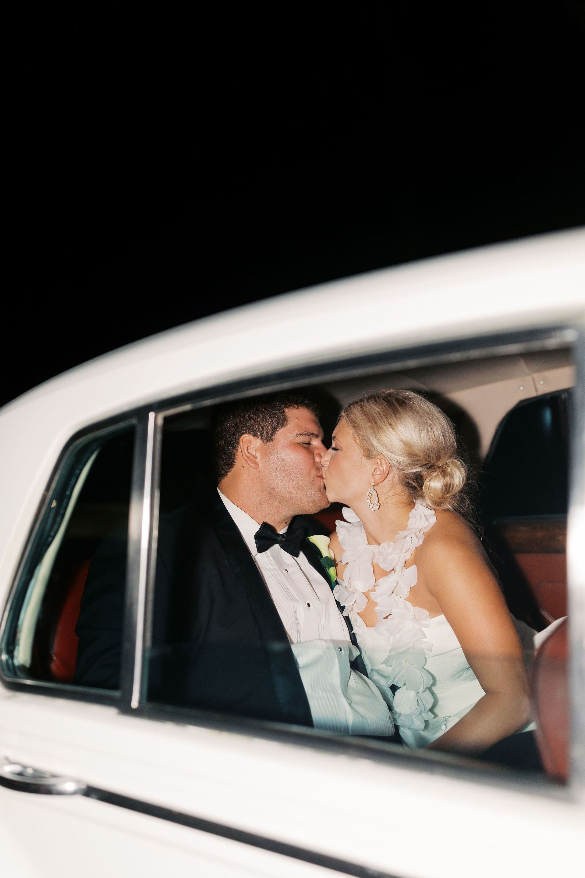 Newlyweds kissing in the back of a white vintage car at night.