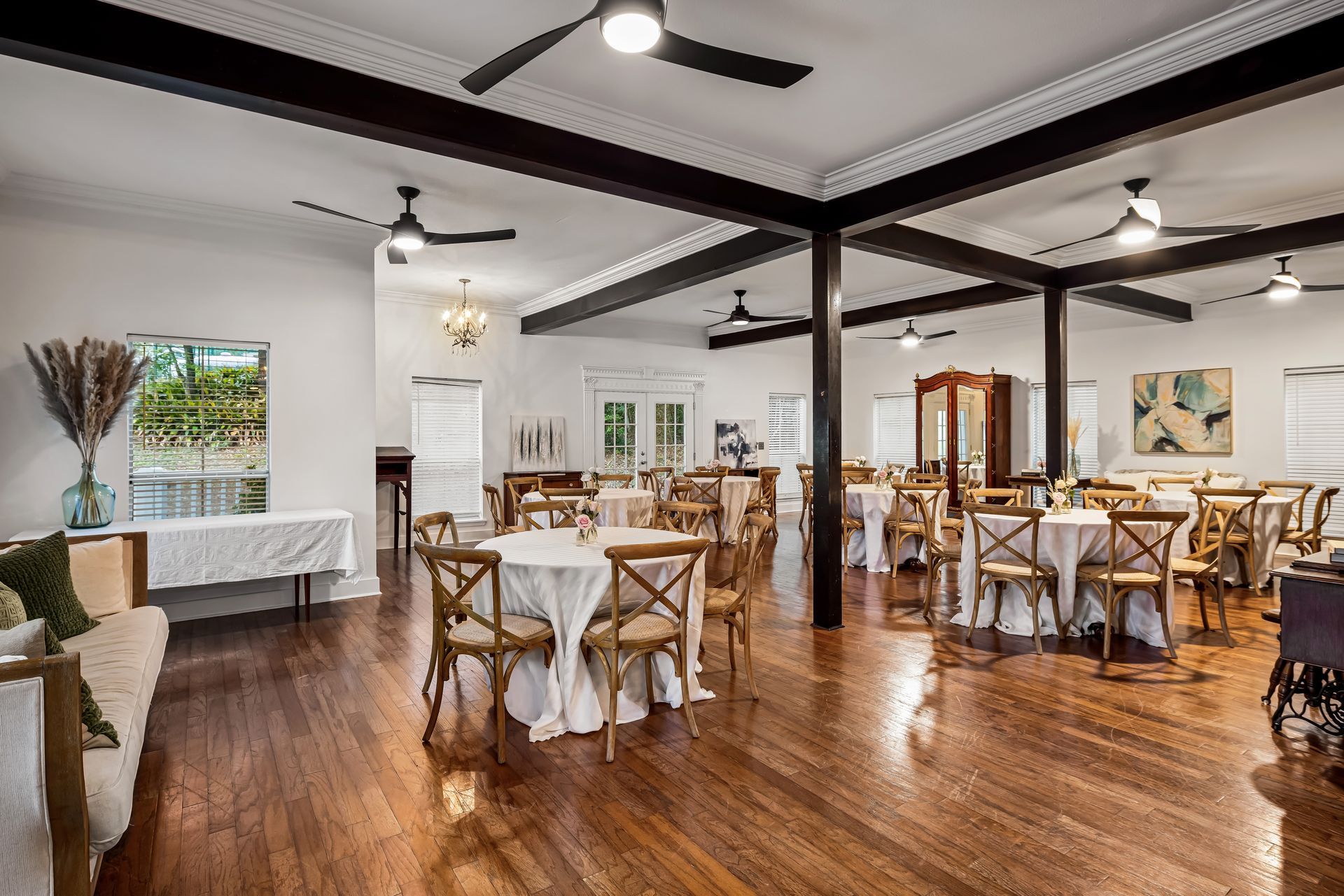 Elegant event space with round tables, wooden chairs, dark beams, and a hardwood floor.