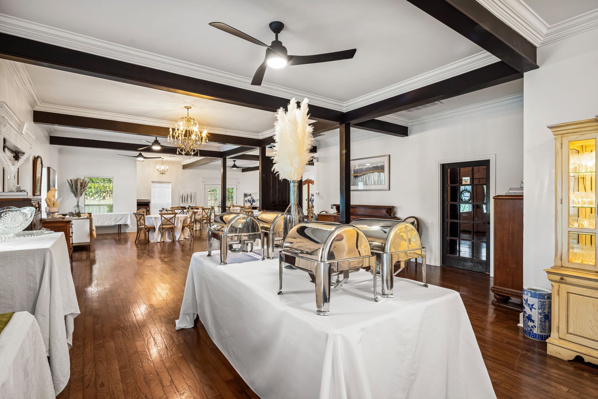 Formal dining room with buffet tables, hardwood floors, chandeliers, and decorative ceiling beams.