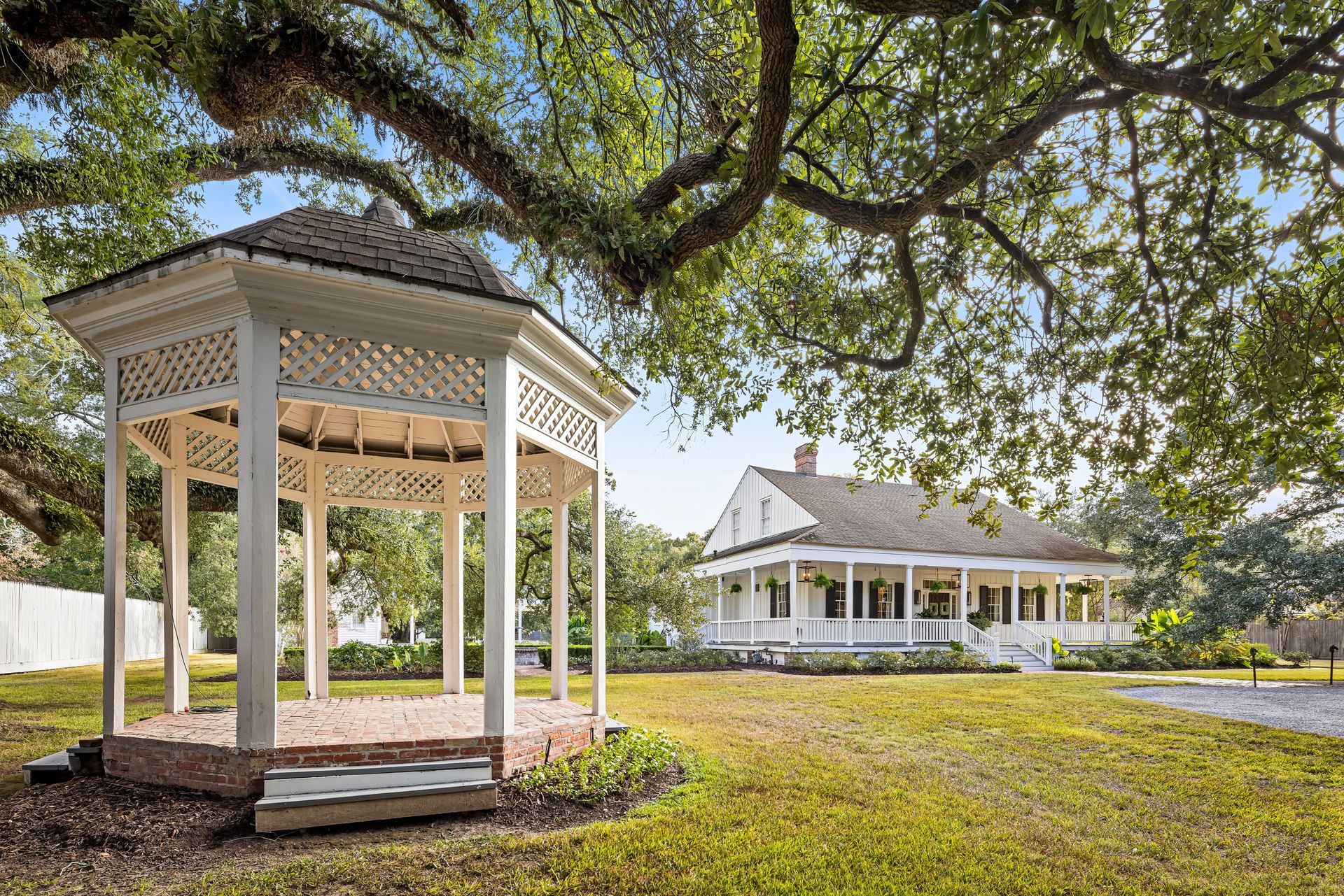 White gazebo in a grassy yard, with a white house in the background beneath a large tree.