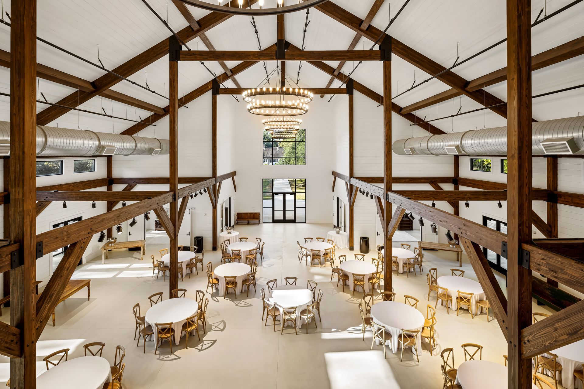 Large open event space with wood beams, round tables set for a gathering, and chandeliers.