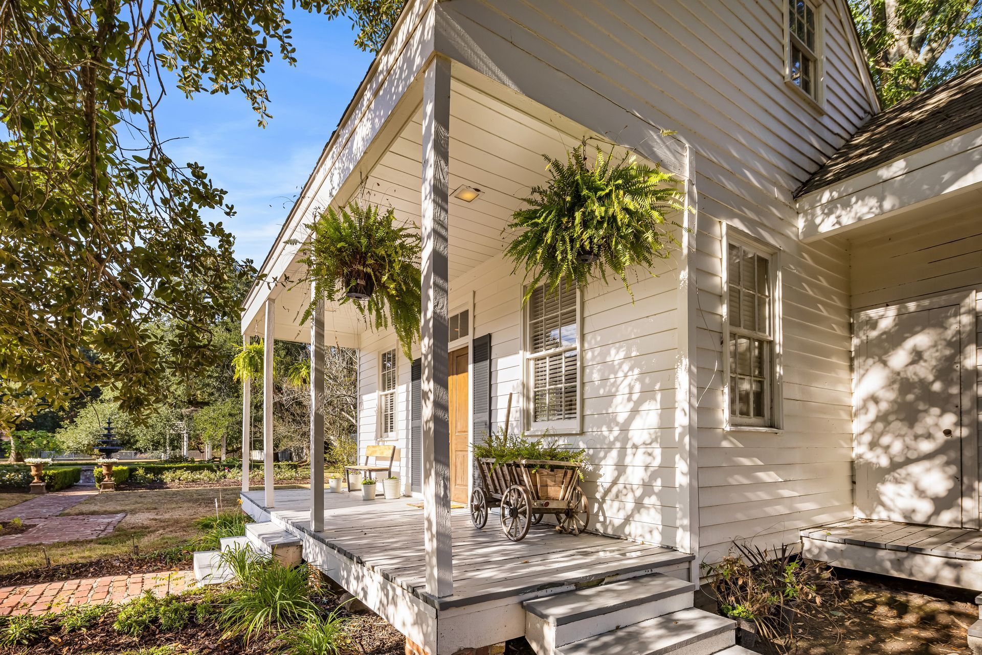 White house with porch, hanging ferns, and brick path in a sunny setting.