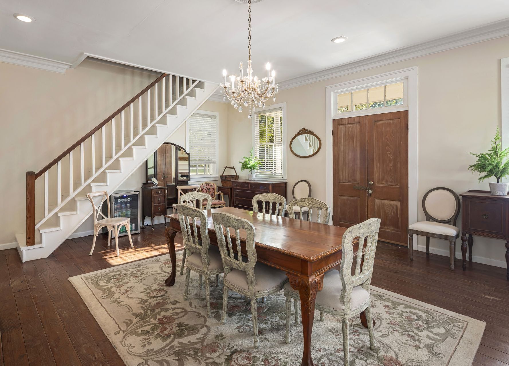Dining room with table, chairs, chandelier, and a staircase. Antique furniture on dark wood floor.
