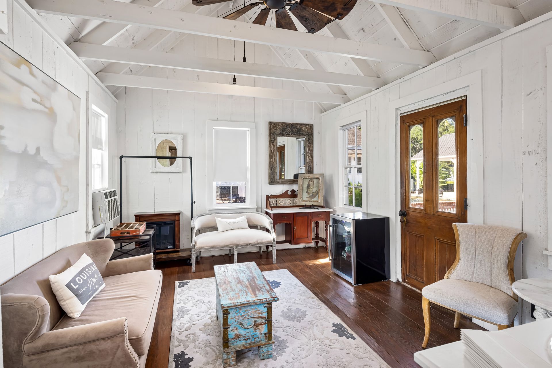 Cozy white-walled living room with wood floors, tan sofa, antique decor, and a wooden door.