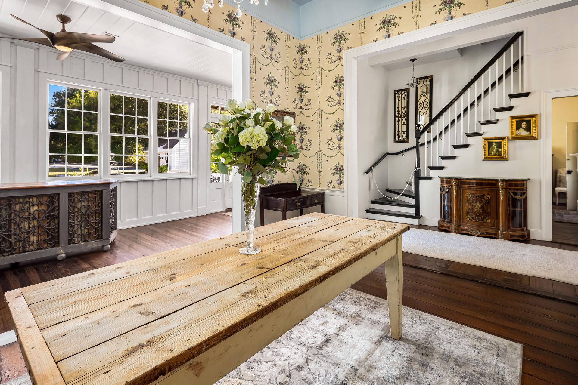 Interior of a home with a wooden table, staircase, floral wallpaper, and large windows.